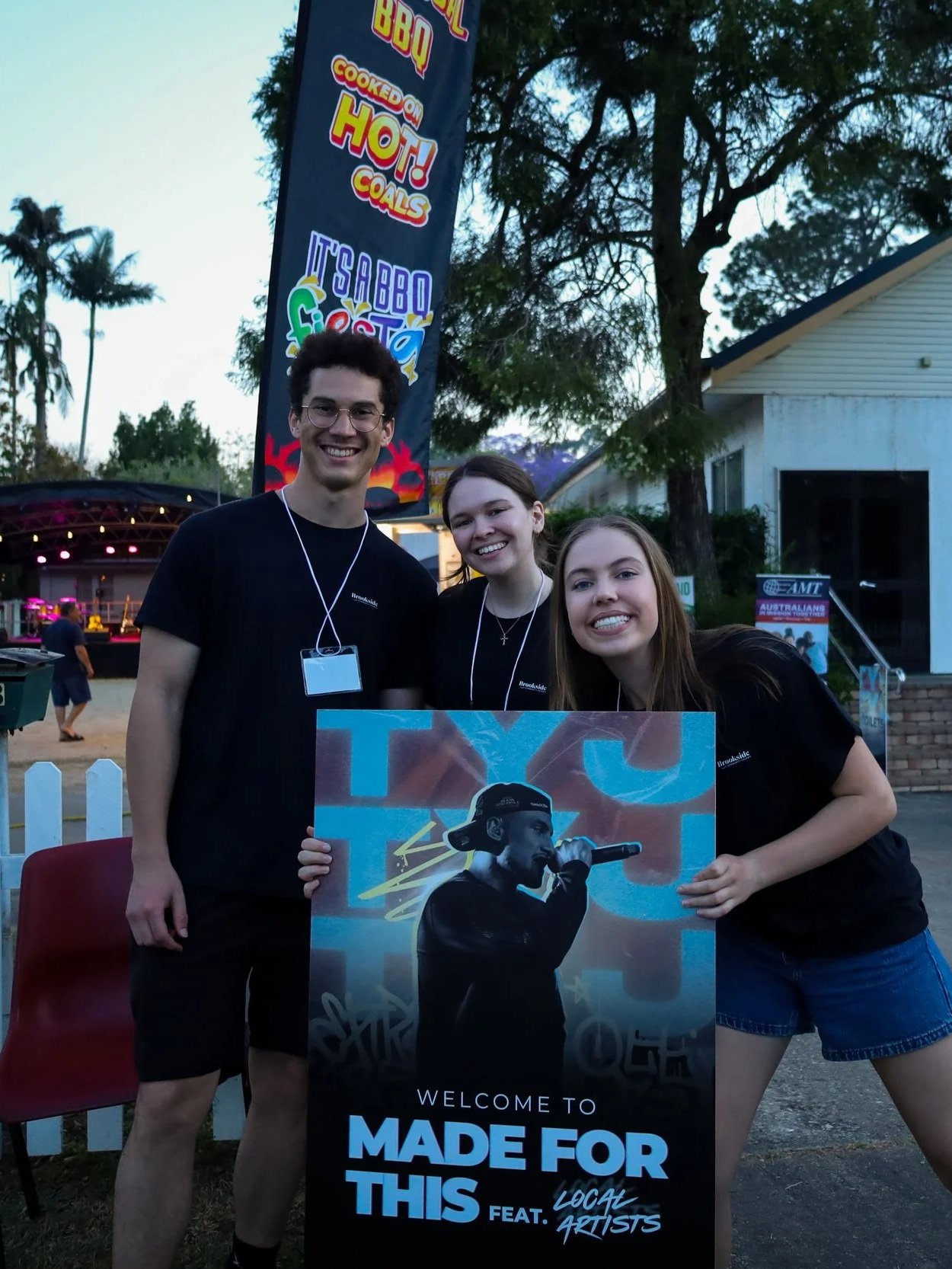 Three young people, two women and one man, are smiling and holding a sign at an outdoor event. The sign features a male performer singing into a microphone with graffiti-style background. The event banner in the background promotes a BBQ festival with text about hot coals and a BBQ fest.