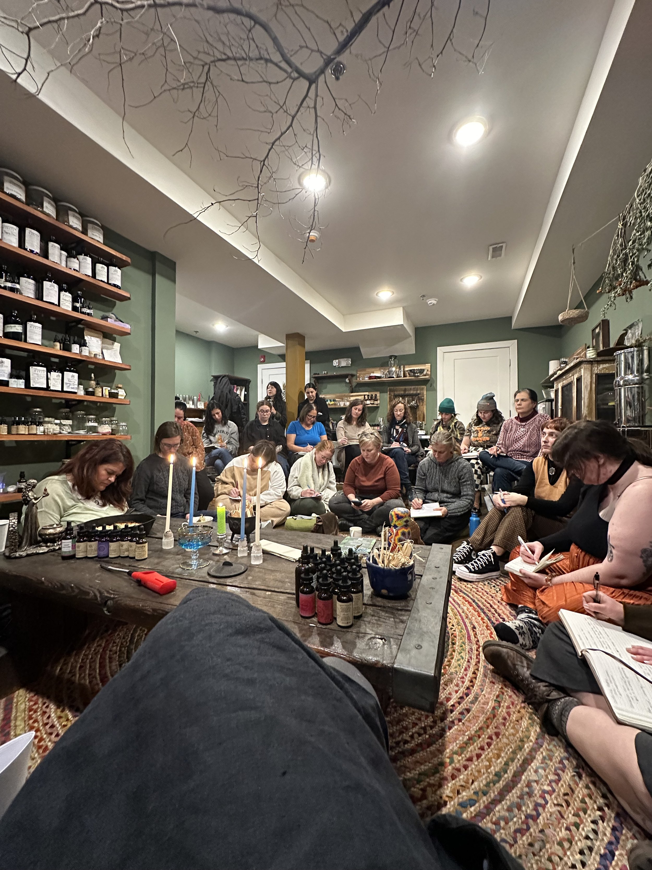People sitting cross-legged on the floor in a cozy, dimly lit room, attending a workshop or seminar with notebooks and pens, with candles and bottles placed on a low table in the center, and shelves with various jars and containers in the background.