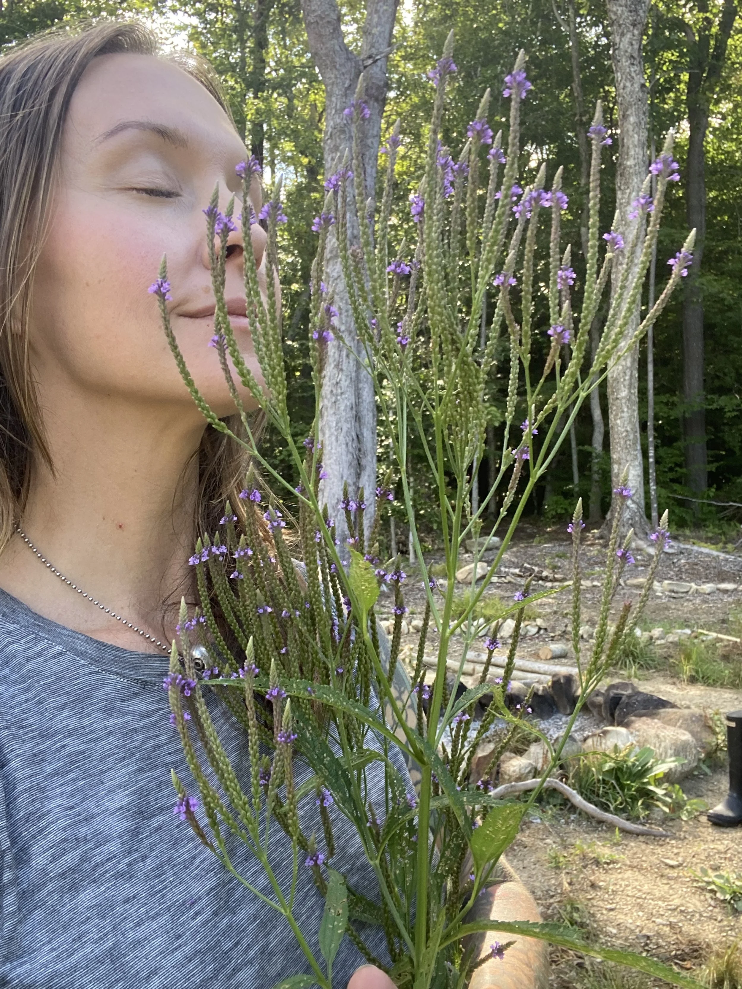 A woman with closed eyes smelling purple wildflowers in a forested outdoor setting.