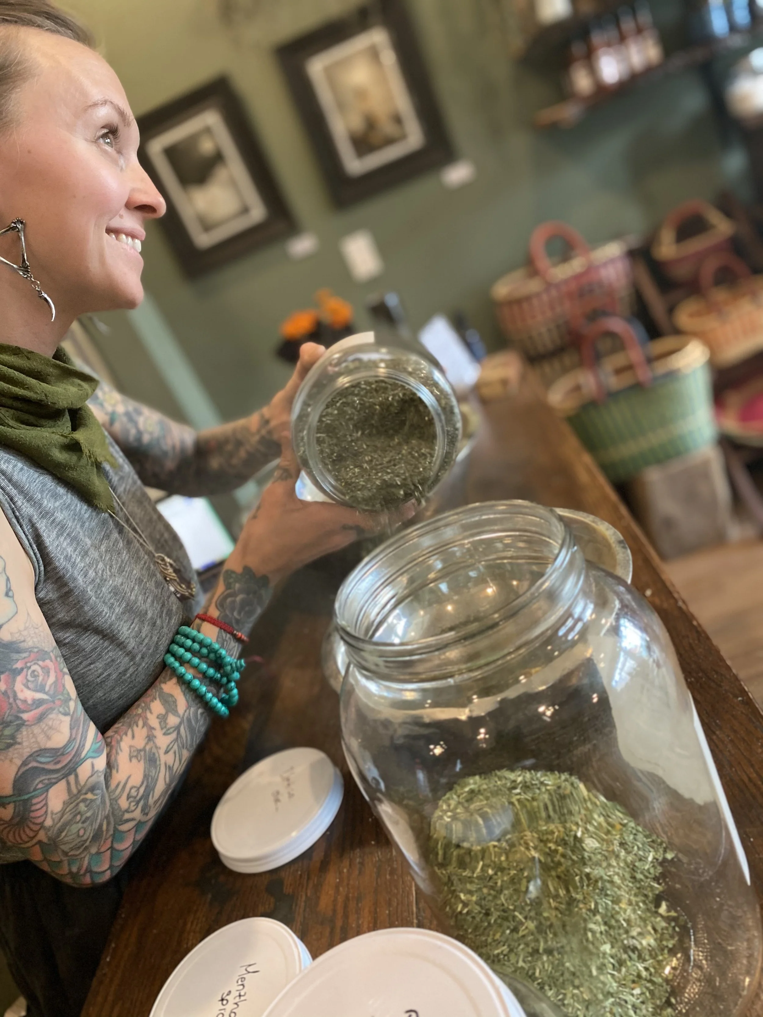 A woman with tattoos and colorful beads on her wrist is smiling and holding a jar of dried herbs in a cozy store or workshop. There is a large glass jar of dried herbs on the table, with other jars and baskets in the background.