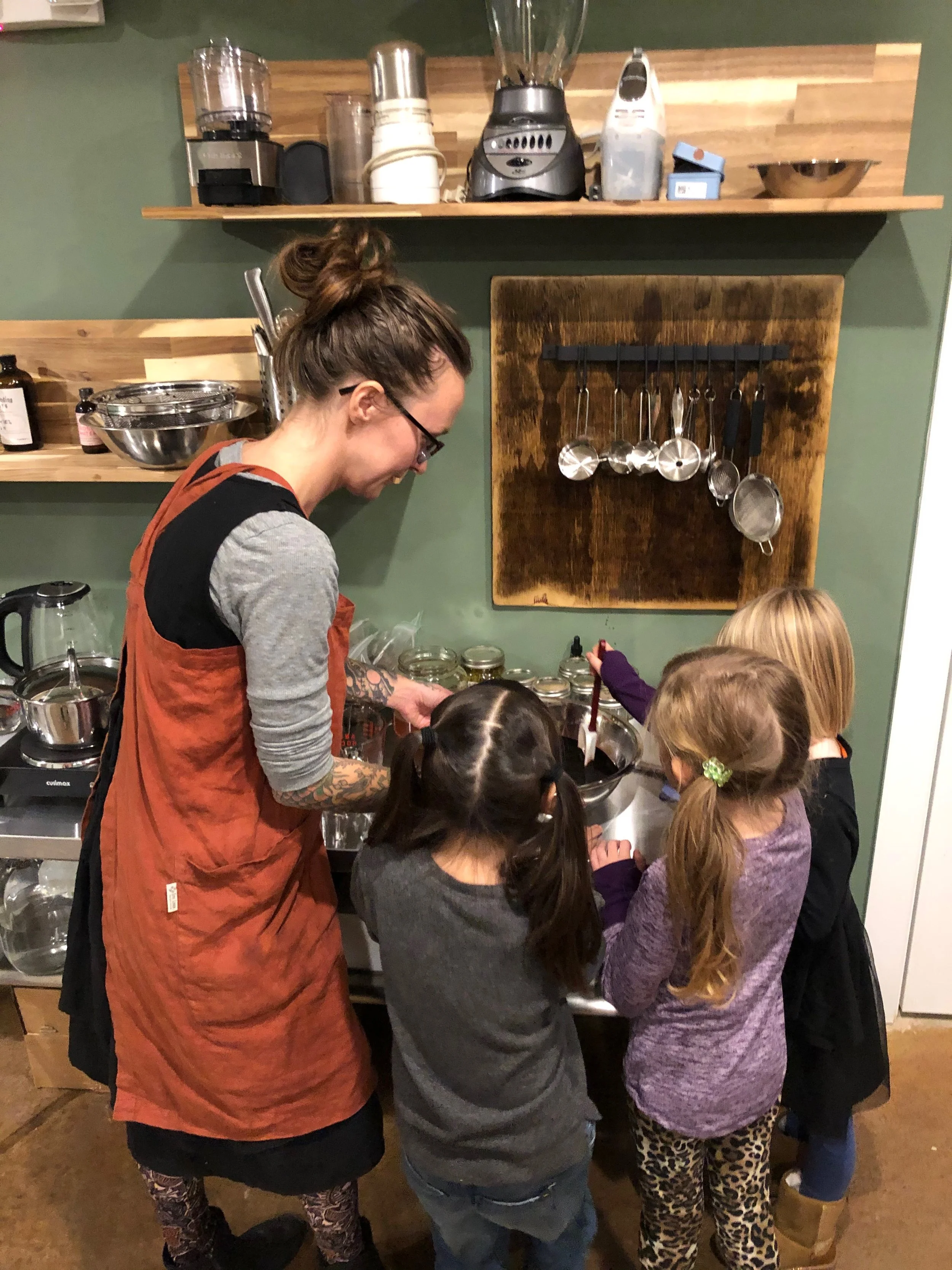 A woman and three children preparing food in a kitchen with wooden shelves and green walls.