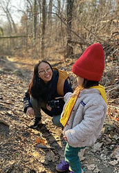 A woman crouching and smiling at a young girl outdoors on a wooded trail in autumn, with the woman wearing glasses and a dark jacket, and the girl wearing a red hat and yellow backpack.
