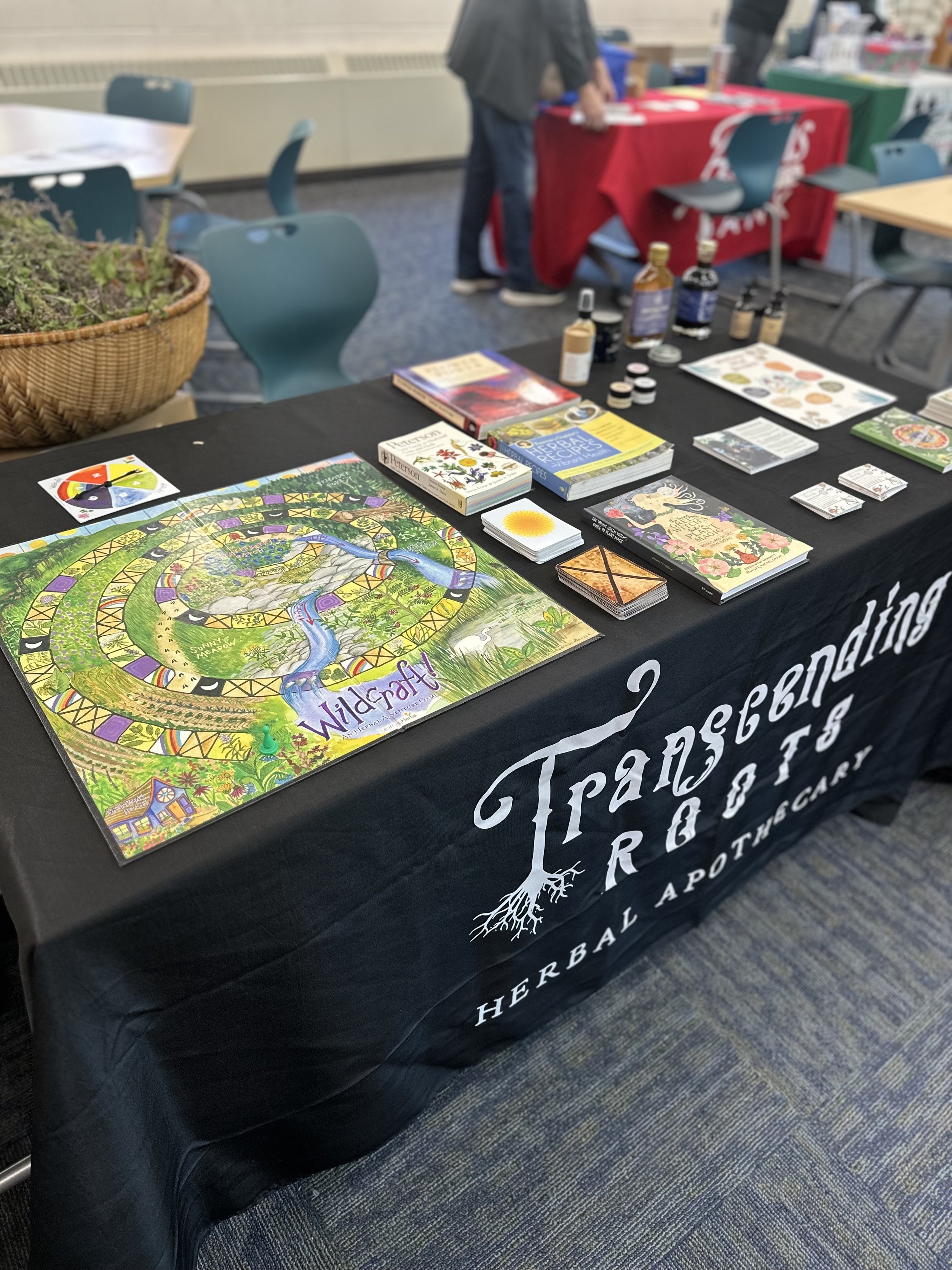 A table display of herbal products and educational materials for herbal gardening, with a black tablecloth printed with 'Transcending Roots Herbal & Pottery'. The table includes a colorful 'Wildcraft' game board, herbal books, small jars of herbal extracts, and informational brochures, with other tables and a plant basket visible in the background.