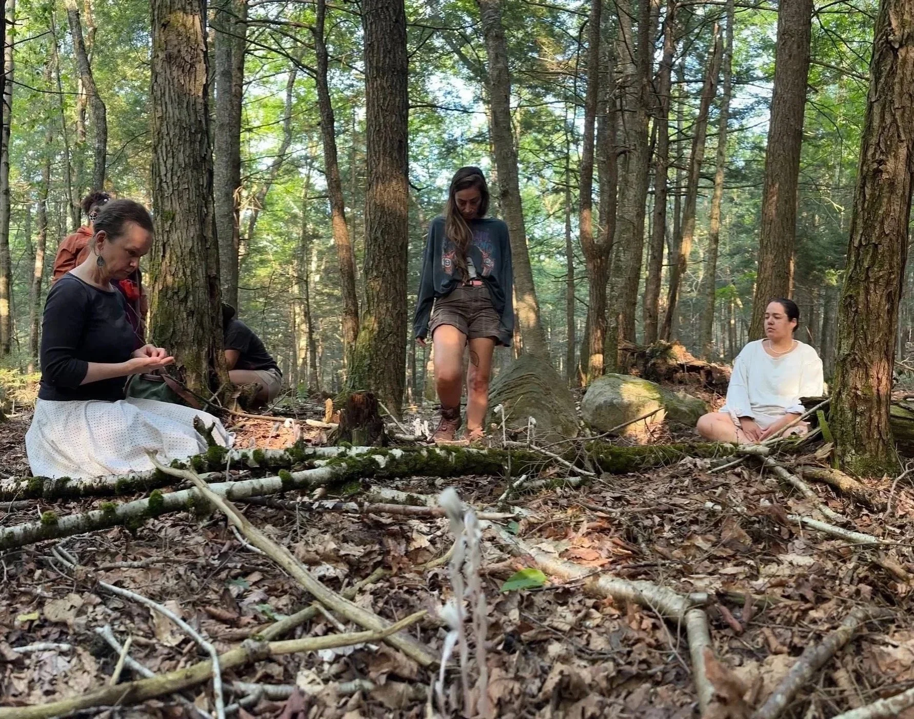 Group of women in a forest, one kneeling and looking down, one standing with her head bowed, and one sitting cross-legged, engaged in a meditative or reflective activity among trees and fallen branches.
