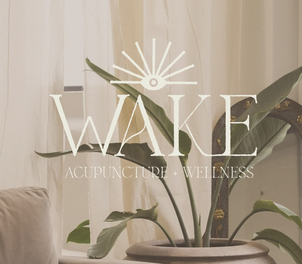 Interior of a wellness space with a large potted plant in the foreground, a mirror with gold accents, and sheer curtains. Overlay text reads 'WAKE ACUPUNCTURE + WELLNESS' with a stylized sun or eye symbol.