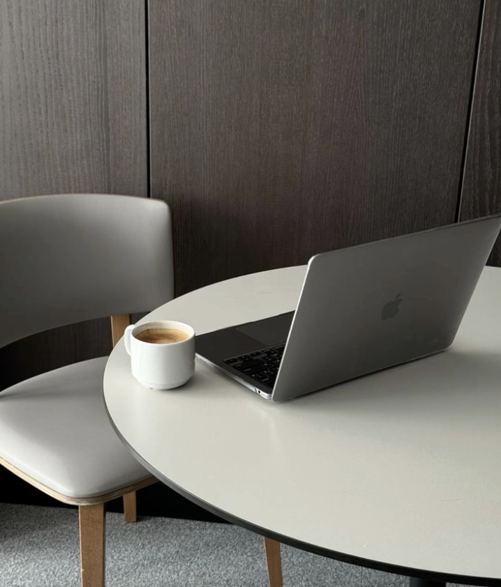 A white round table with a silver laptop and a white coffee mug filled with coffee, next to a gray chair in front of a wood-paneled wall.