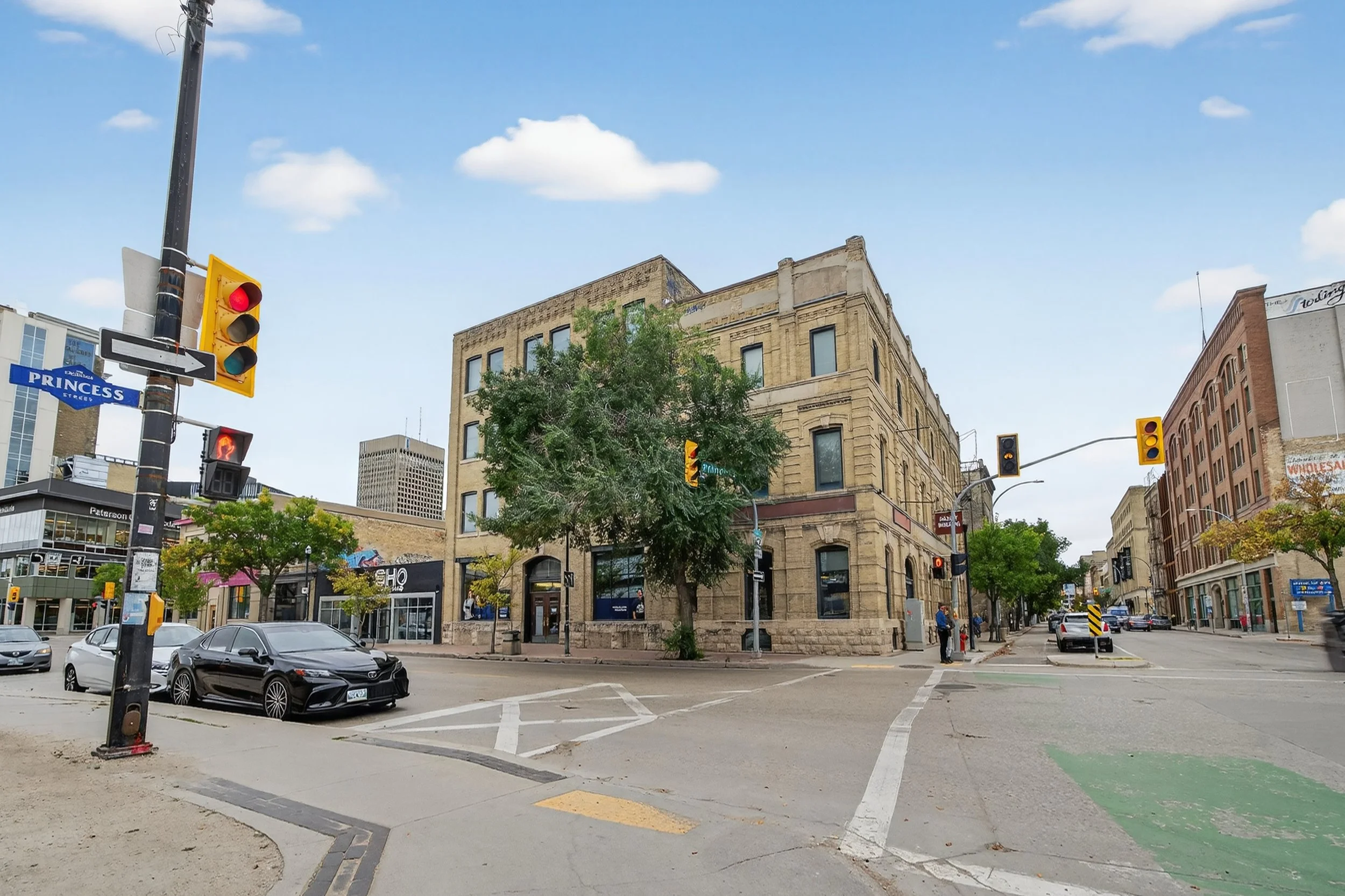 A city street intersection with traffic lights, parked cars, trees, and buildings under a partly cloudy sky.