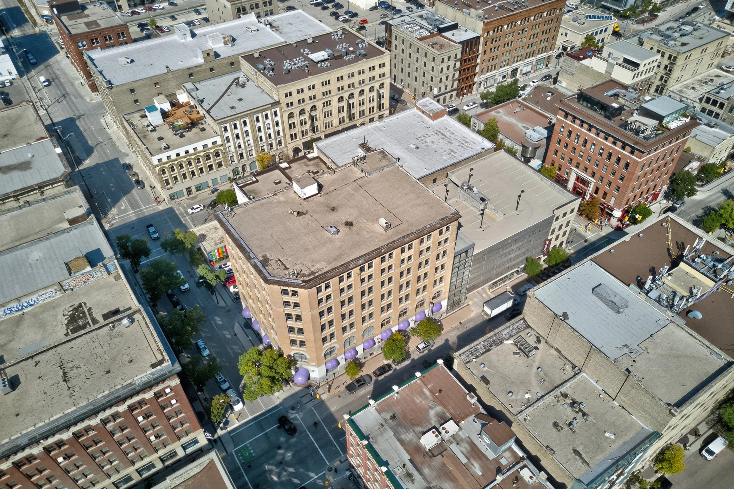 Aerial view of a city block showing several multi-story buildings, streets with parked cars, and some trees.