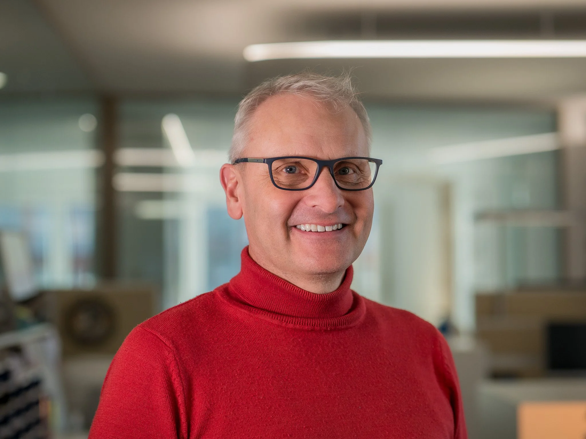 Ein freundlicher älterer Mann mit Brille, trägt einen roten Pullover, lächelt und steht in einem Büro.