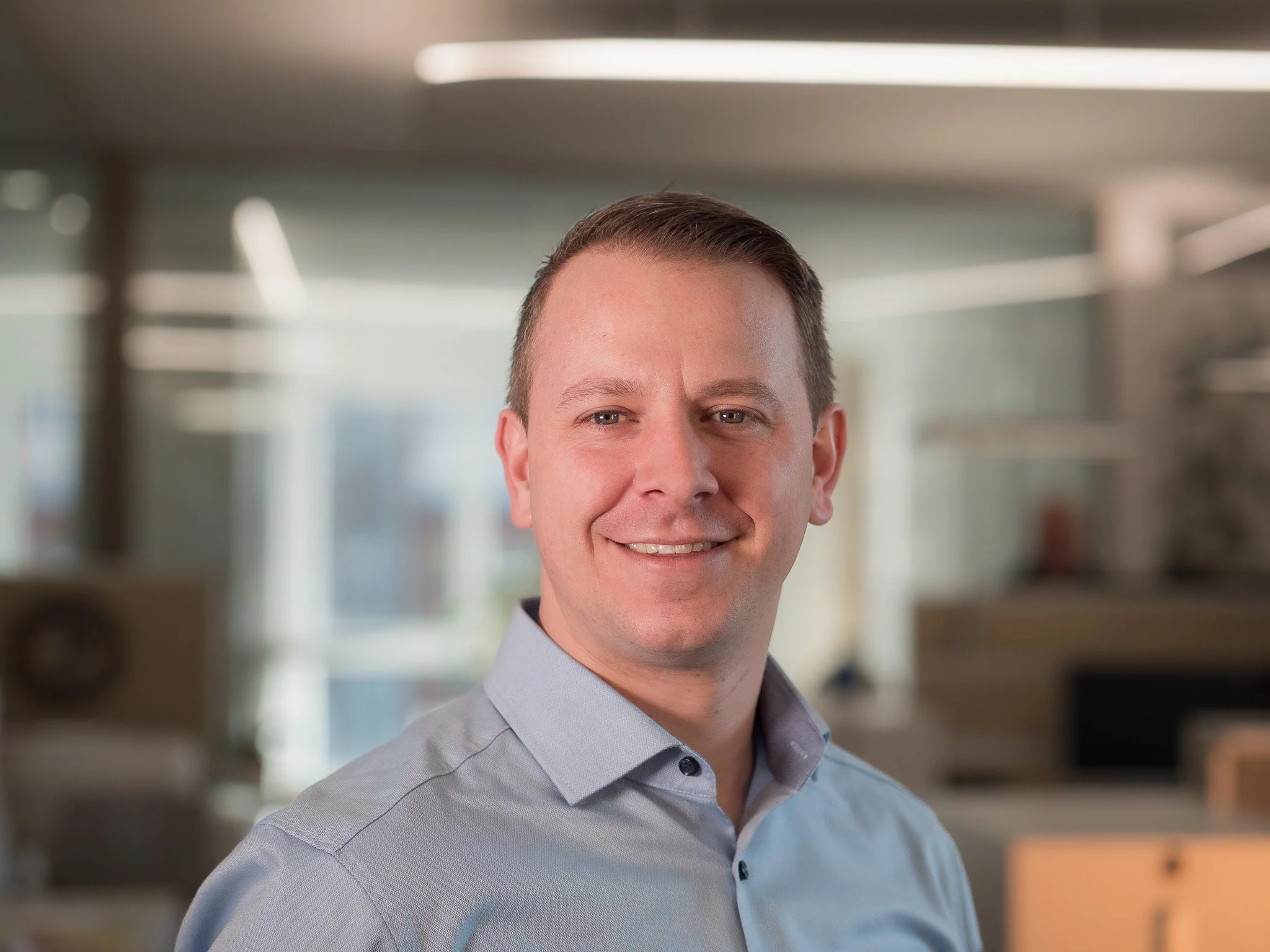 Ein Mann in einem hellblauen Hemd, lächelnd, im Büro mit moderner, lichtdurchfluteter Architektur im Hintergrund.