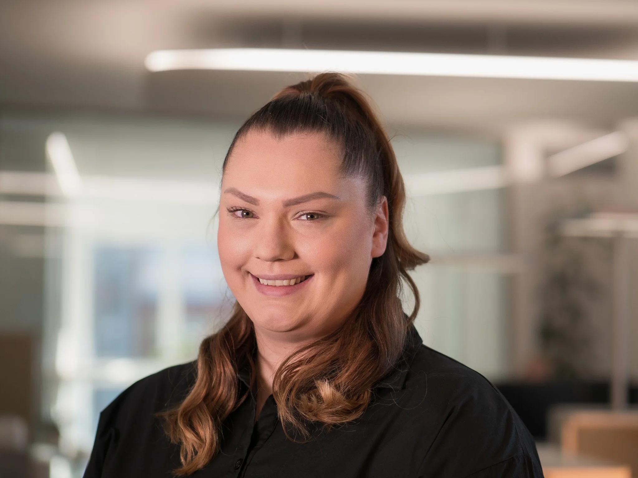 Porträt einer lächelnden Frau mit braunen, gelockten Haaren in einem schwarzen Hemd, im Büro.