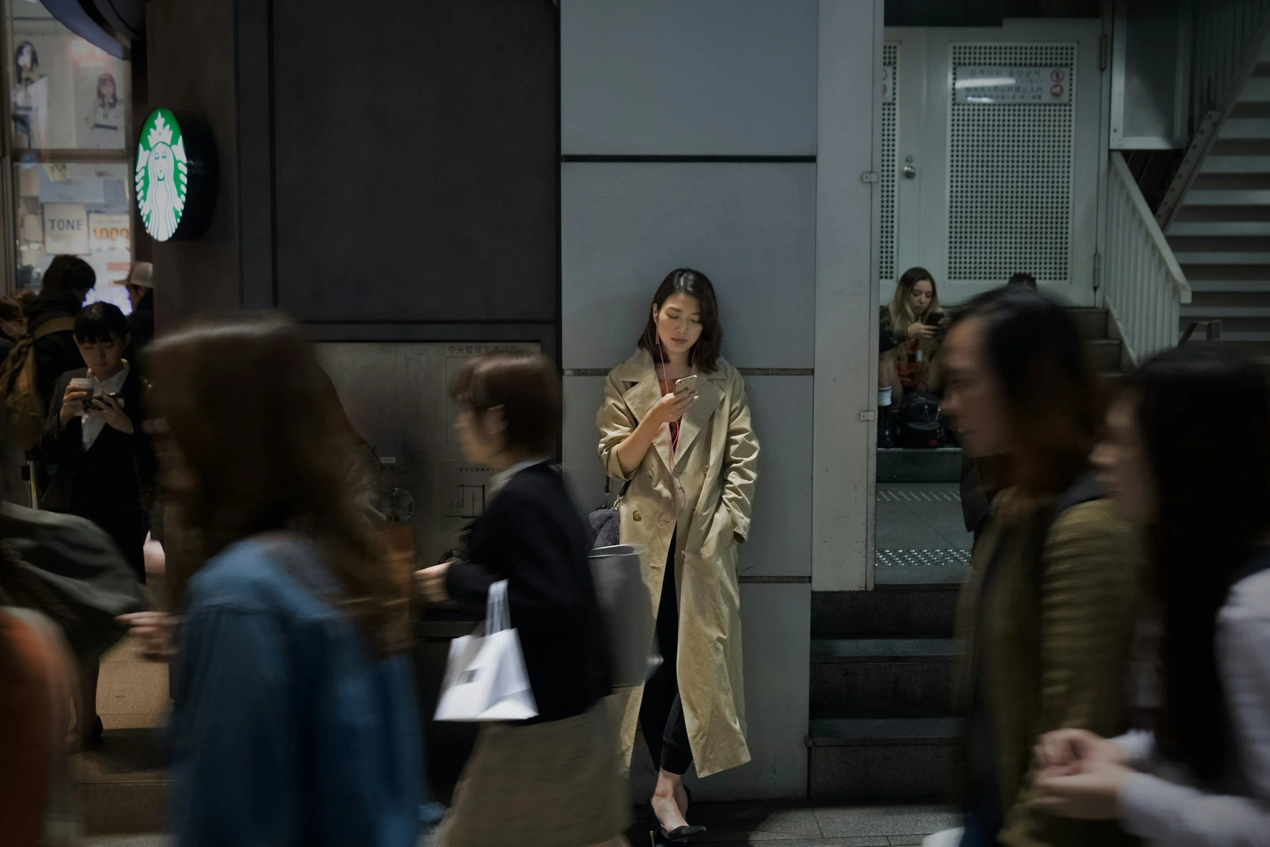 A woman in a beige trench coat standing against a wall in a busy train station, looking at her phone. Several other people are walking by, some also looking at their phones. An illuminated Starbucks sign and staircase are visible in the background.