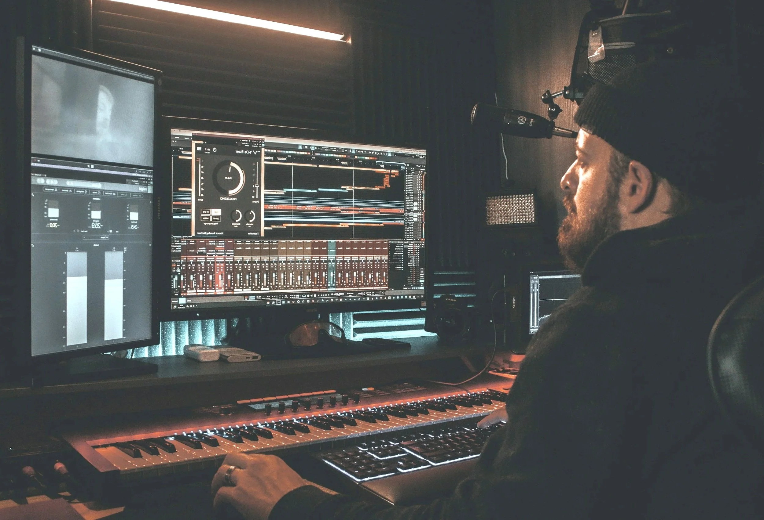 A person working in a music production studio with multiple computer monitors displaying audio editing software, a MIDI keyboard, and studio equipment.