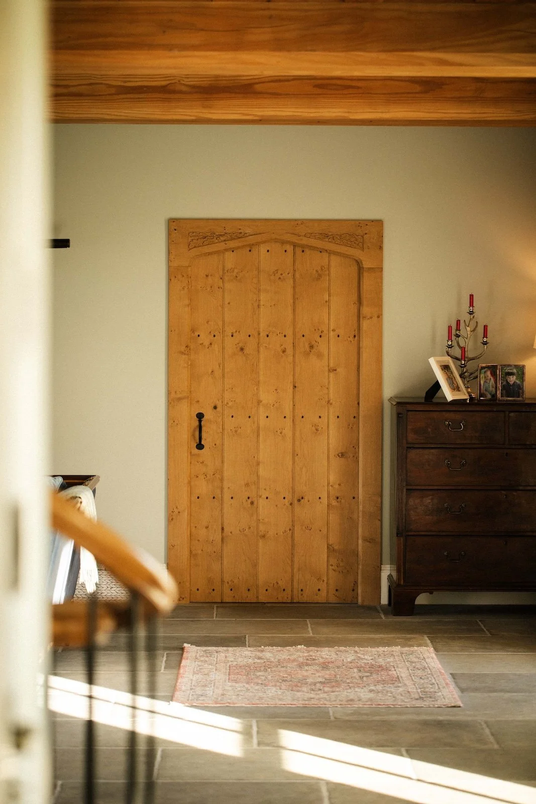 Wooden door with black handle in a room with a dark brown dresser to the right, decorated with framed photos and a candelabra, and a small pinkish rug on the tiled floor.