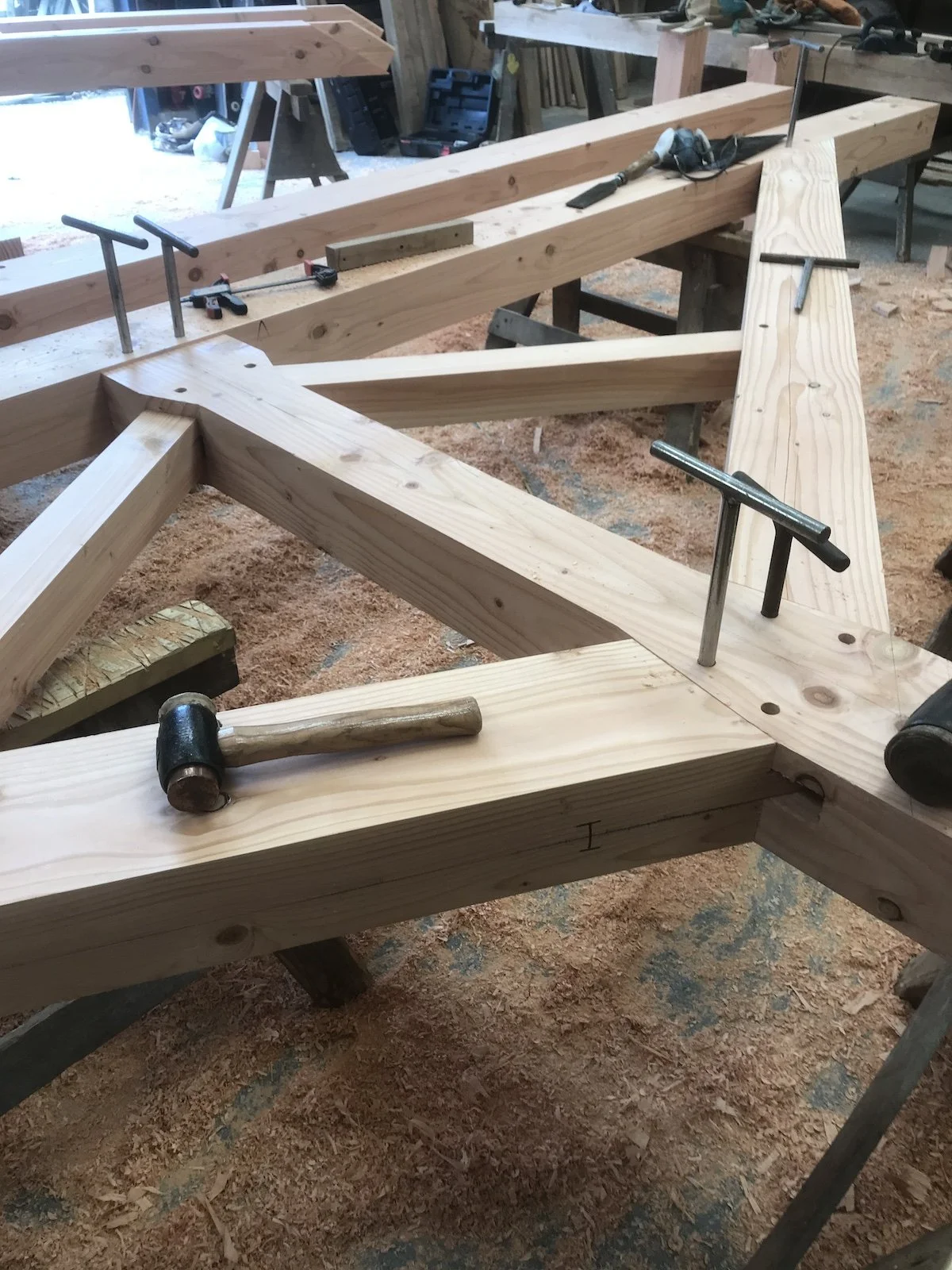 A woodworking workshop with wooden beams, clamps, a hammer, and woodworking tools on a workbench, with sawdust on the floor.