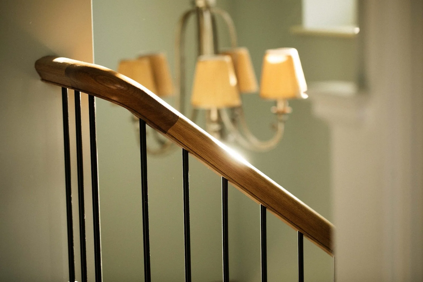 Close-up of a wooden stair handrail with black metal balusters and a blurry background of a wall-mounted lamp fixture with multiple lampshades.