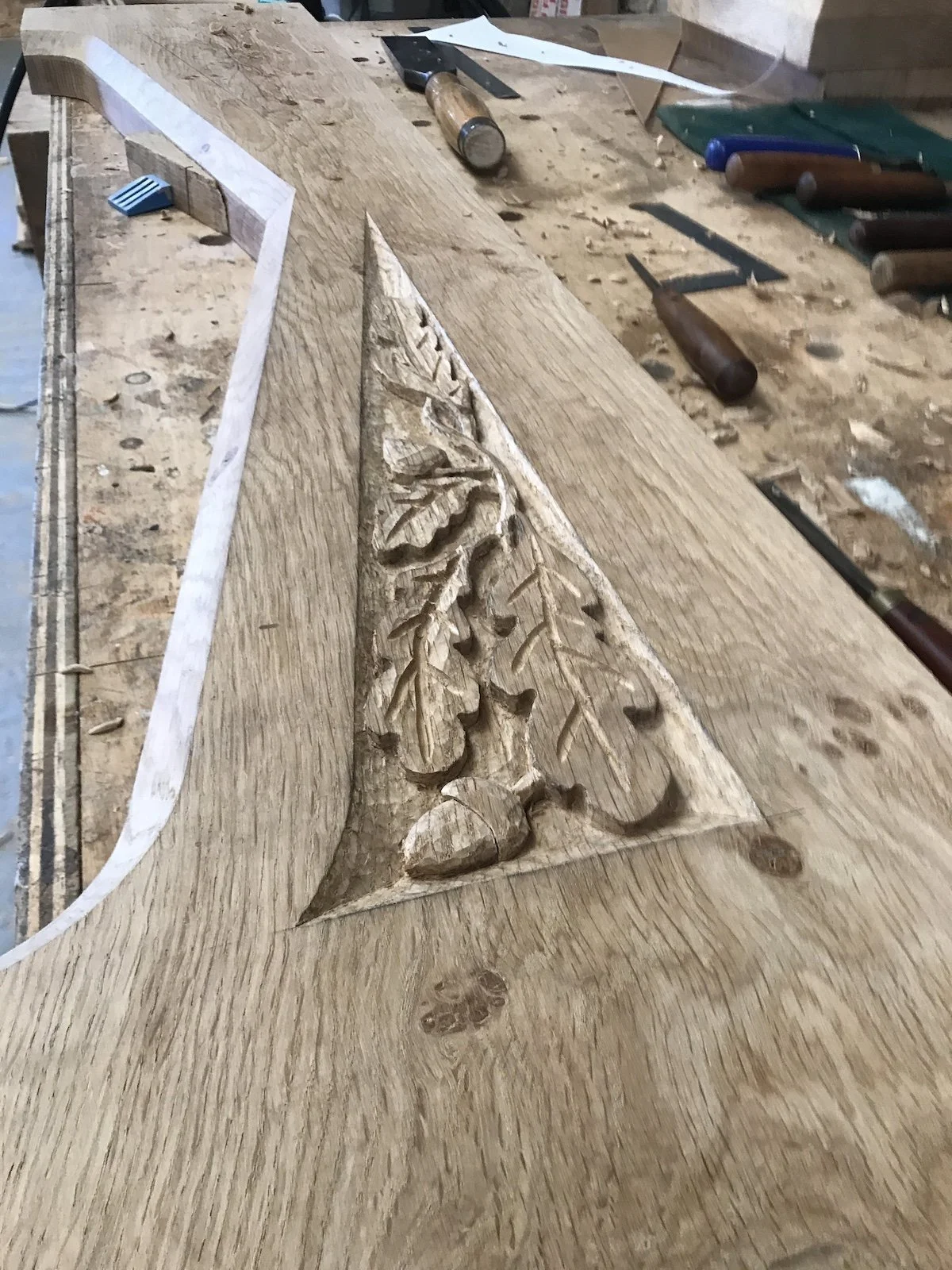 Woodworking in progress with carved leaf pattern on a wooden piece on a workbench surrounded by woodworking tools.