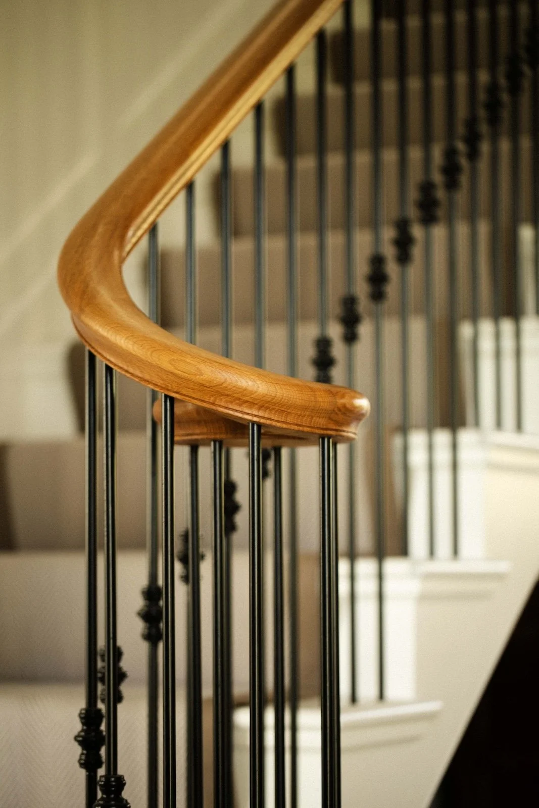 Close-up of a curved wooden handrail on a staircase, with black metal balusters and a beige wall in the background.