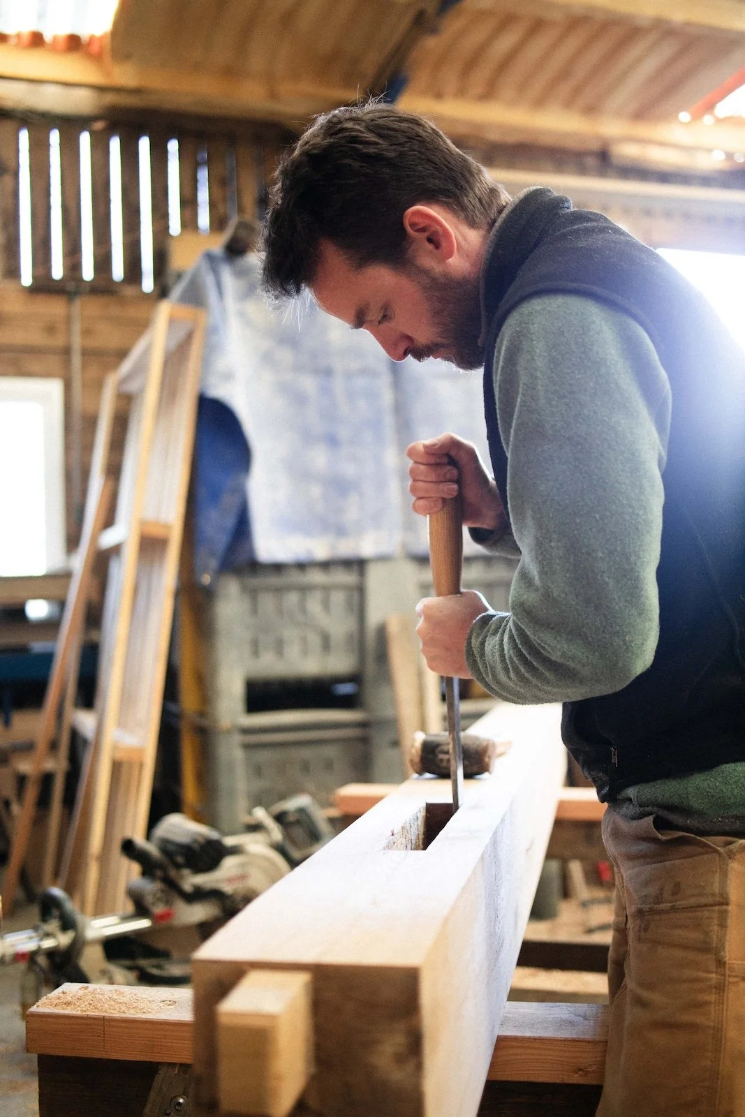 A man with dark hair and a beard, wearing a gray and dark-colored jacket, is working in a woodworking shop, using a chisel and hammer on a long piece of wood. The workshop has a rustic wooden interior with various tools and materials in the background.