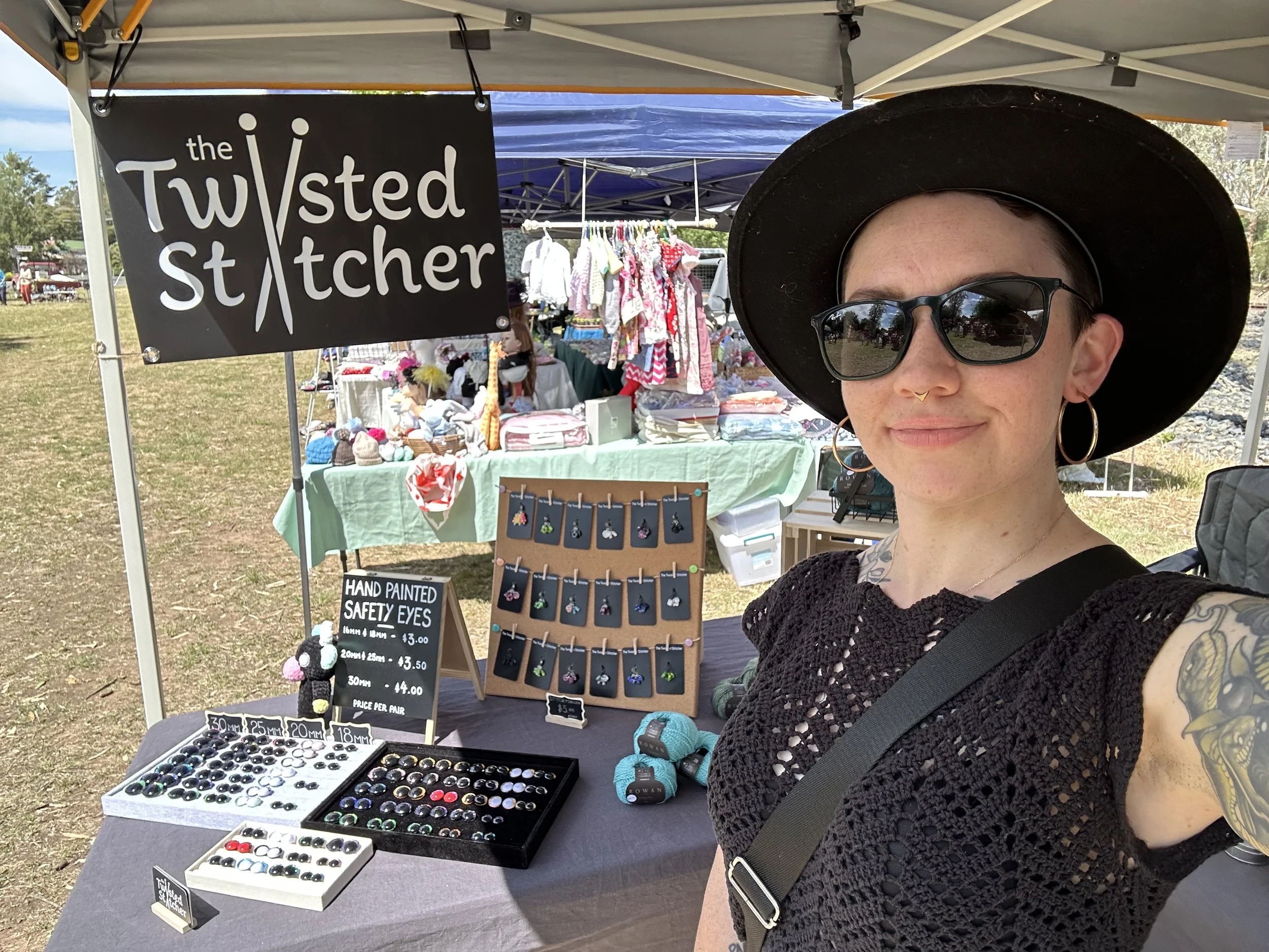 A woman wearing a black hat, sunglasses, and earrings is taking a selfie at an outdoor craft market, which displays handmade safety eyes.