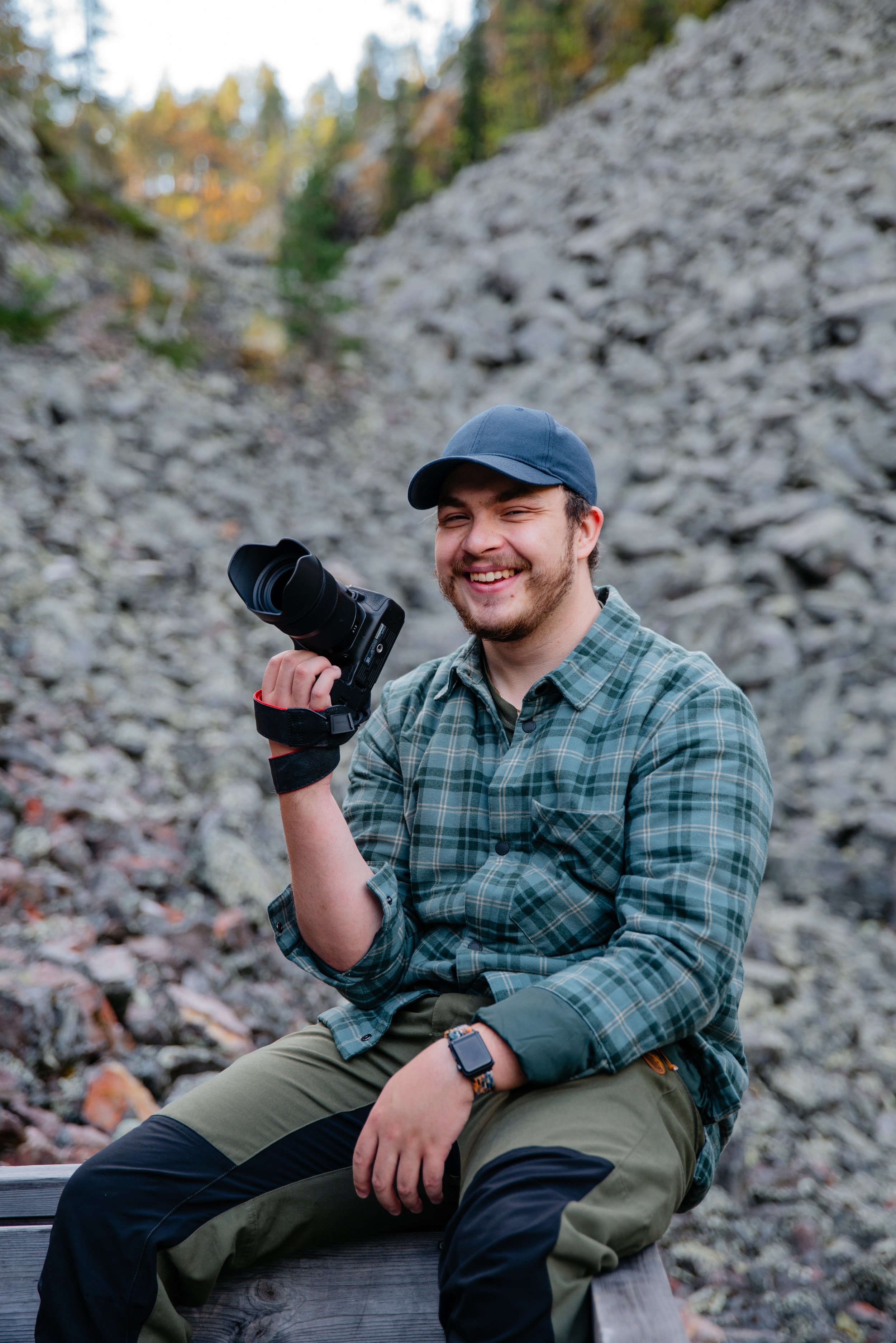 A man sitting outdoors on a wooden bench, smiling and holding a camera, with rocky terrain and trees in the background.
