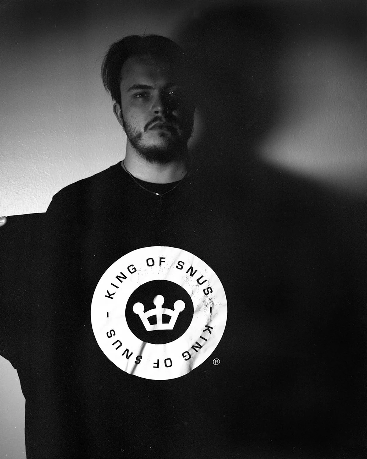 A young man with dark hair and a beard wearing a black shirt with a "King of Snus" logo on it, standing against a plain wall, with his face partly shadowed.