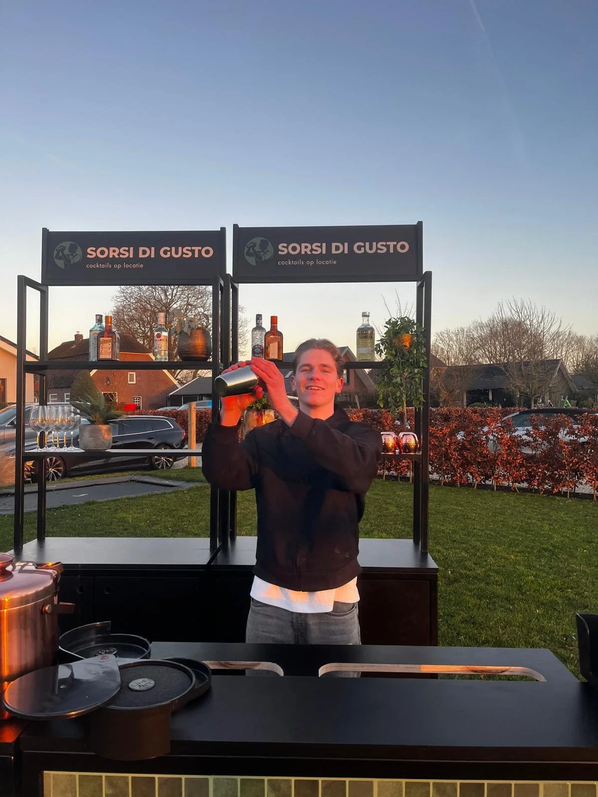 Bartender achter mobiele bar met cocktailshaker, achtergrond met cocktailbarelementen, logo 'Sorsi di Gusto'.