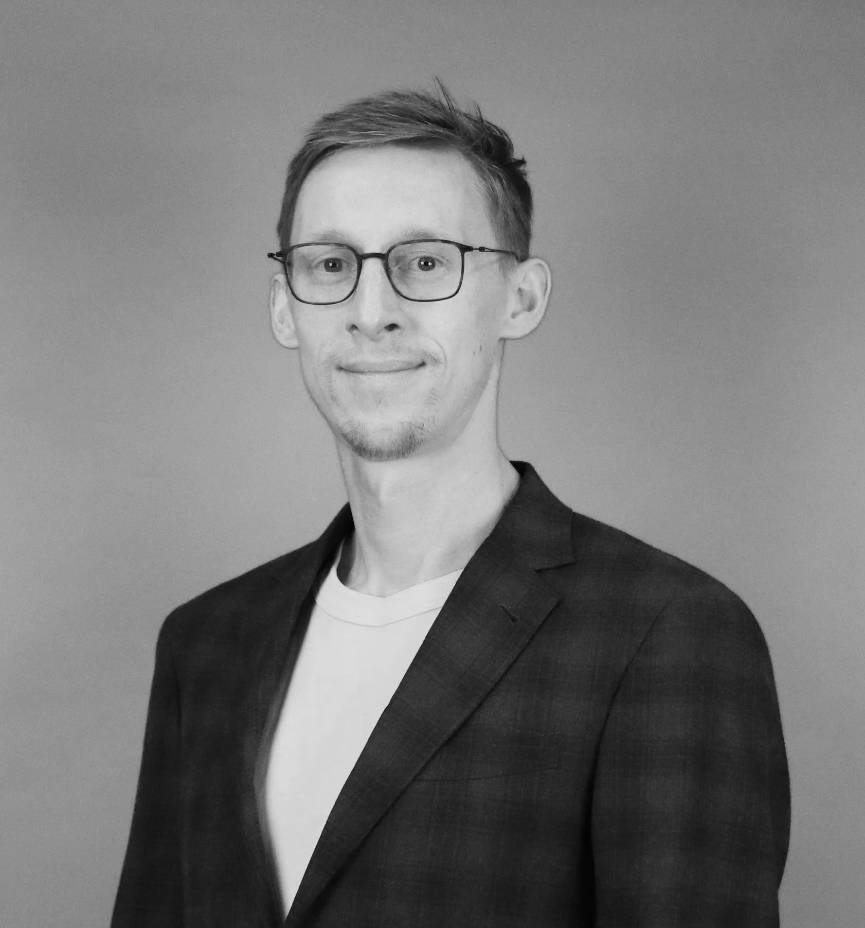 Black and white portrait of a man with glasses, short hair, wearing a suit jacket over a t-shirt, smiling slightly.