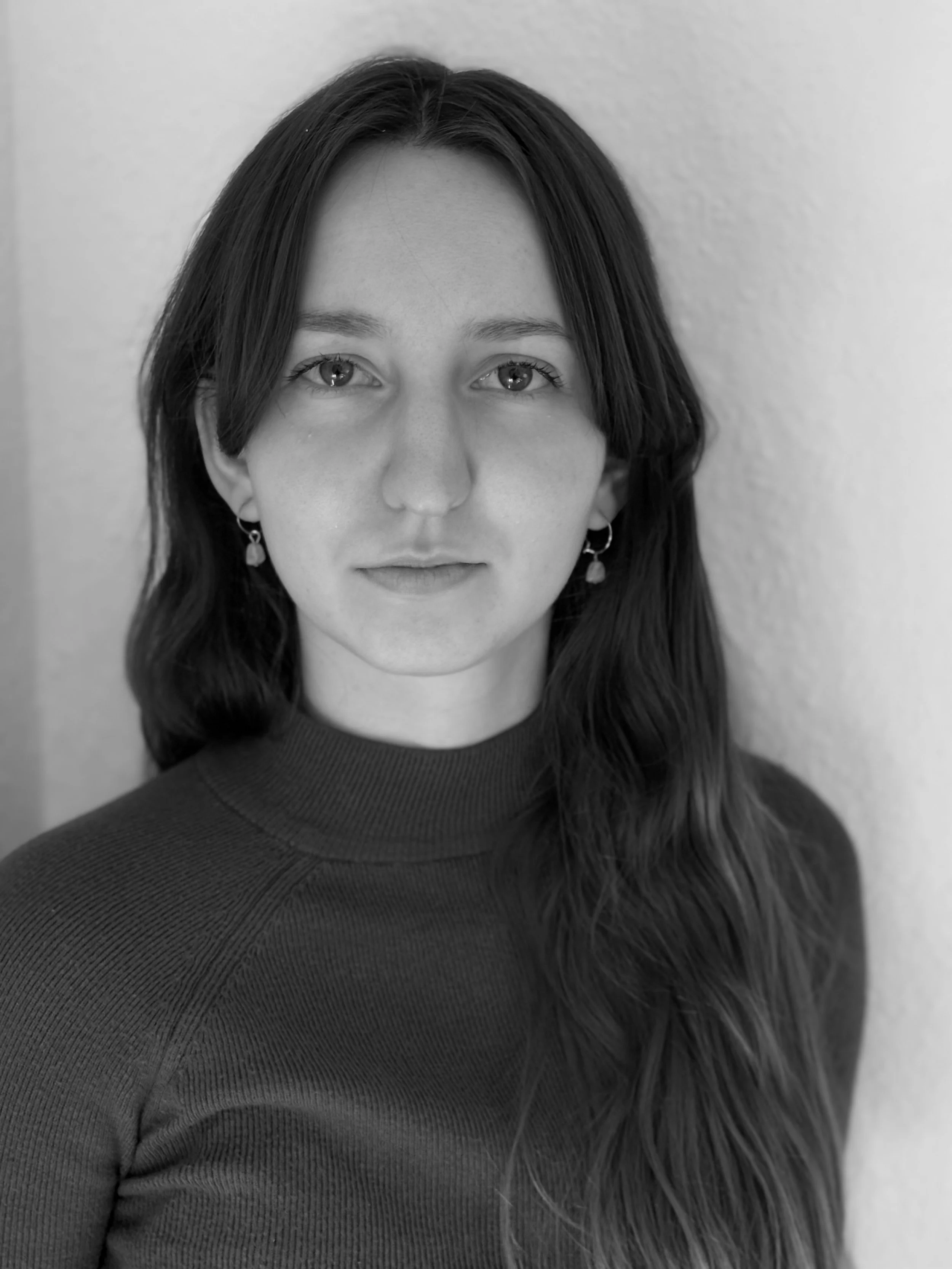 Black and white portrait of a young woman with long hair, wearing earrings and a turtleneck sweater, standing against a plain wall.