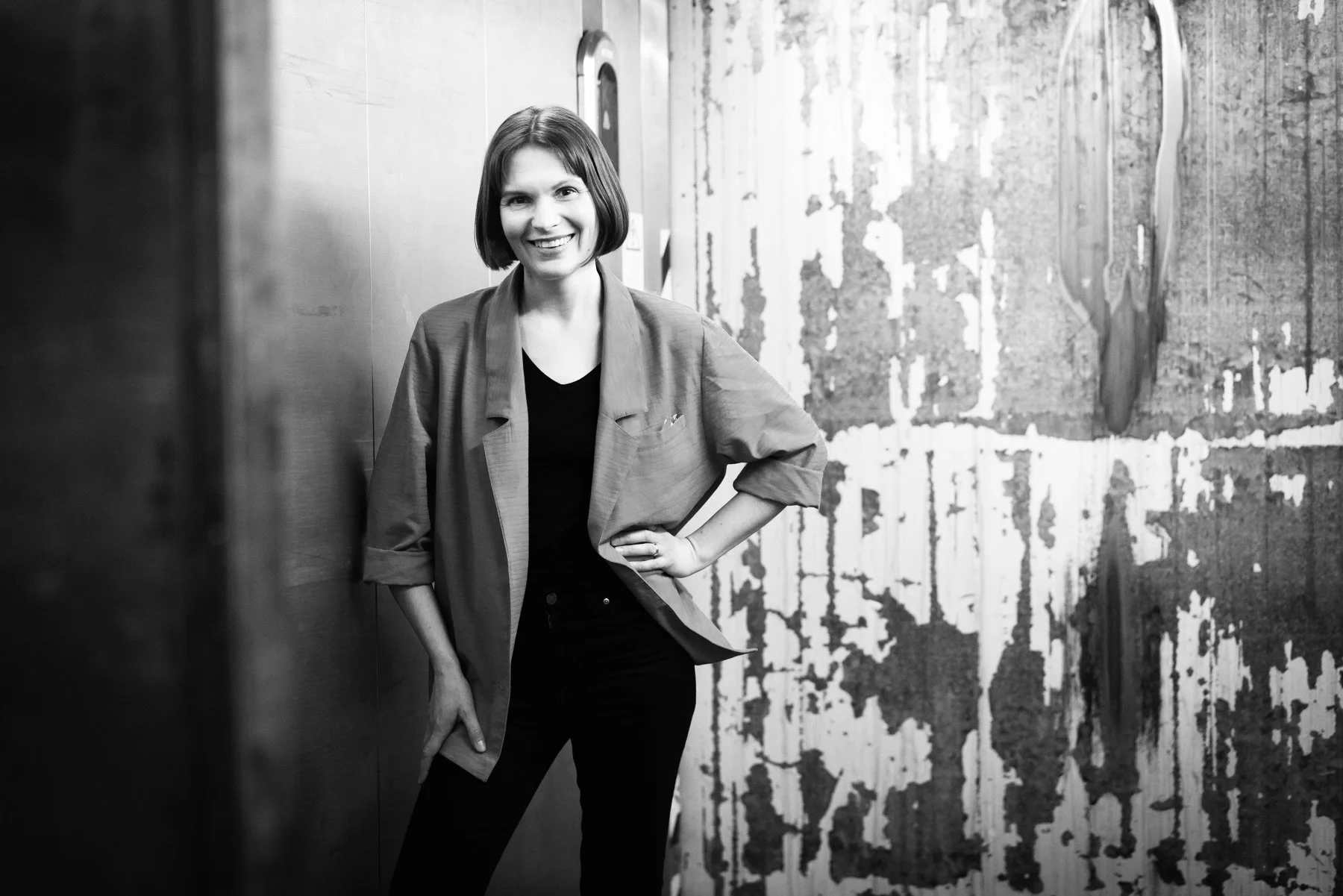 A woman with shoulder-length hair smiling, wearing a blazer over a black top, standing with one hand on her hip against a textured wall in a black and white photo.