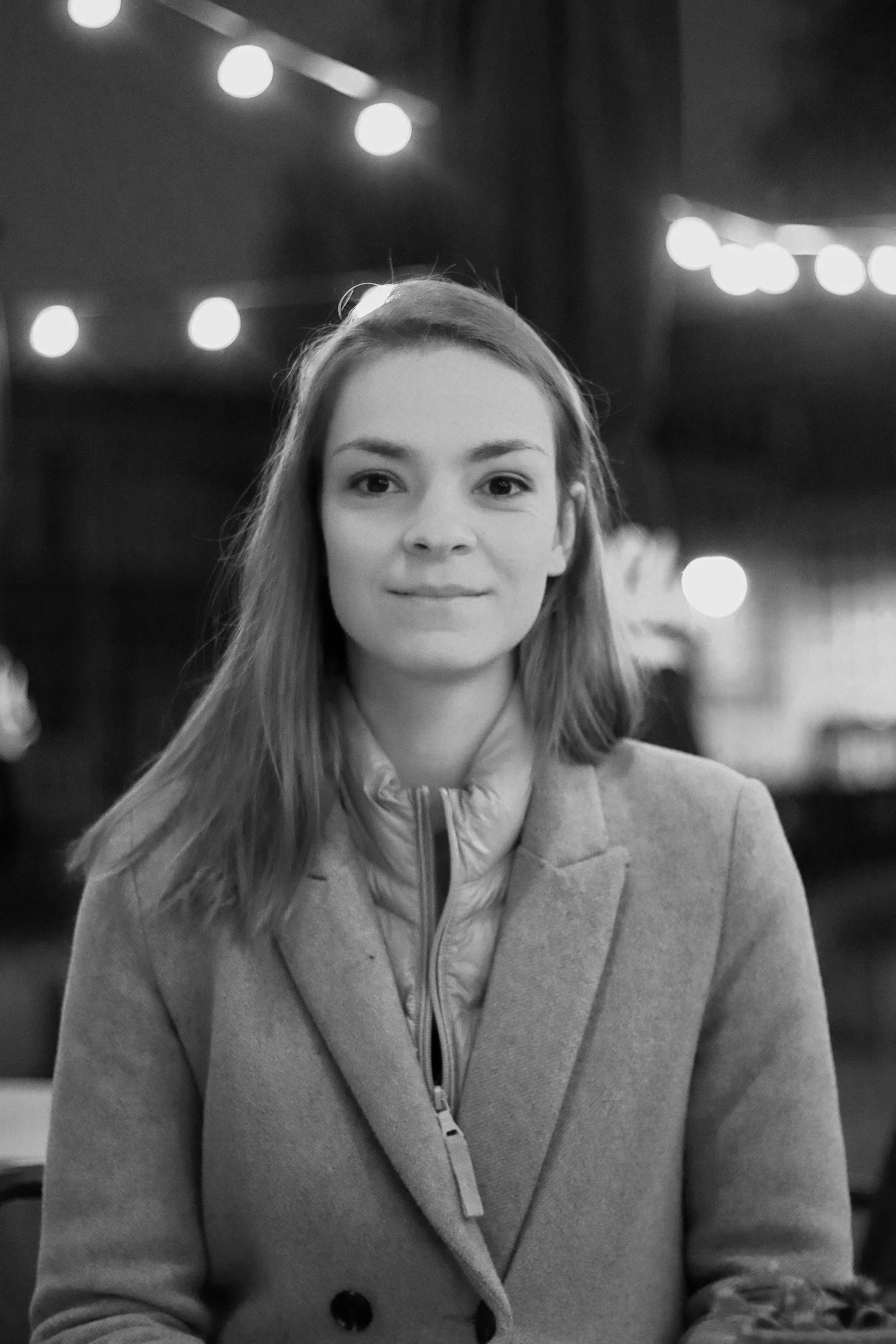 A young woman with long hair, smiling slightly, wearing a coat, in a setting with string lights in the background, captured in black and white.