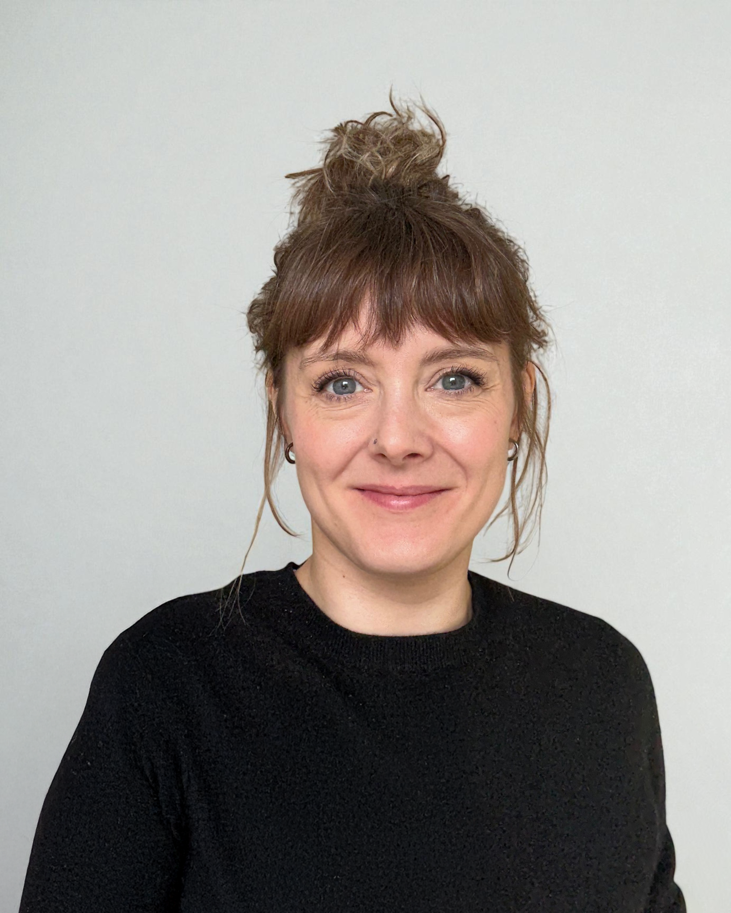 A woman with brown hair styled in a messy bun, wearing a black top, smiling in front of a plain white wall.