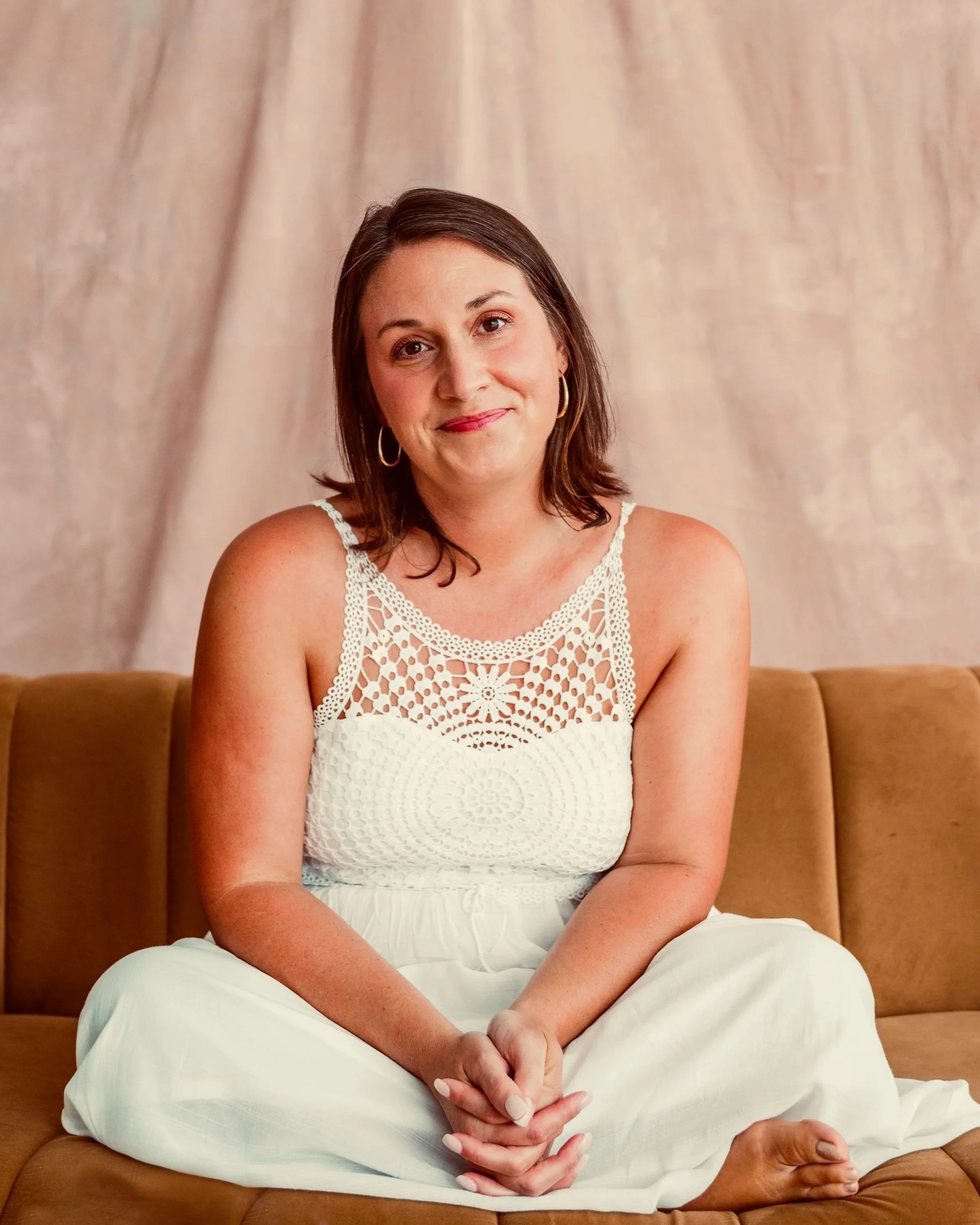 A woman with shoulder-length brown hair, wearing a white crochet top and white pants, sitting cross-legged on a brown sofa against a beige backdrop, smiling softly at the camera.