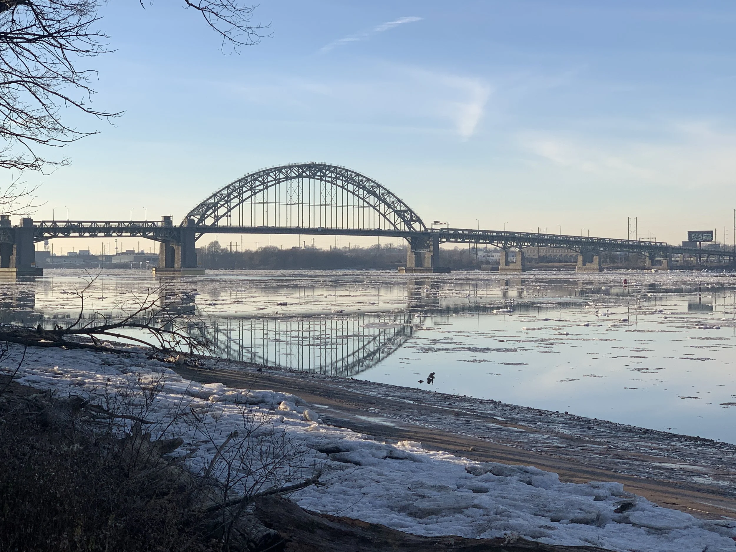 Tacony Palmyra Bridge spans across the Delaware river, with patches of ice floating on the water surface, under a clear blue sky during winter.
