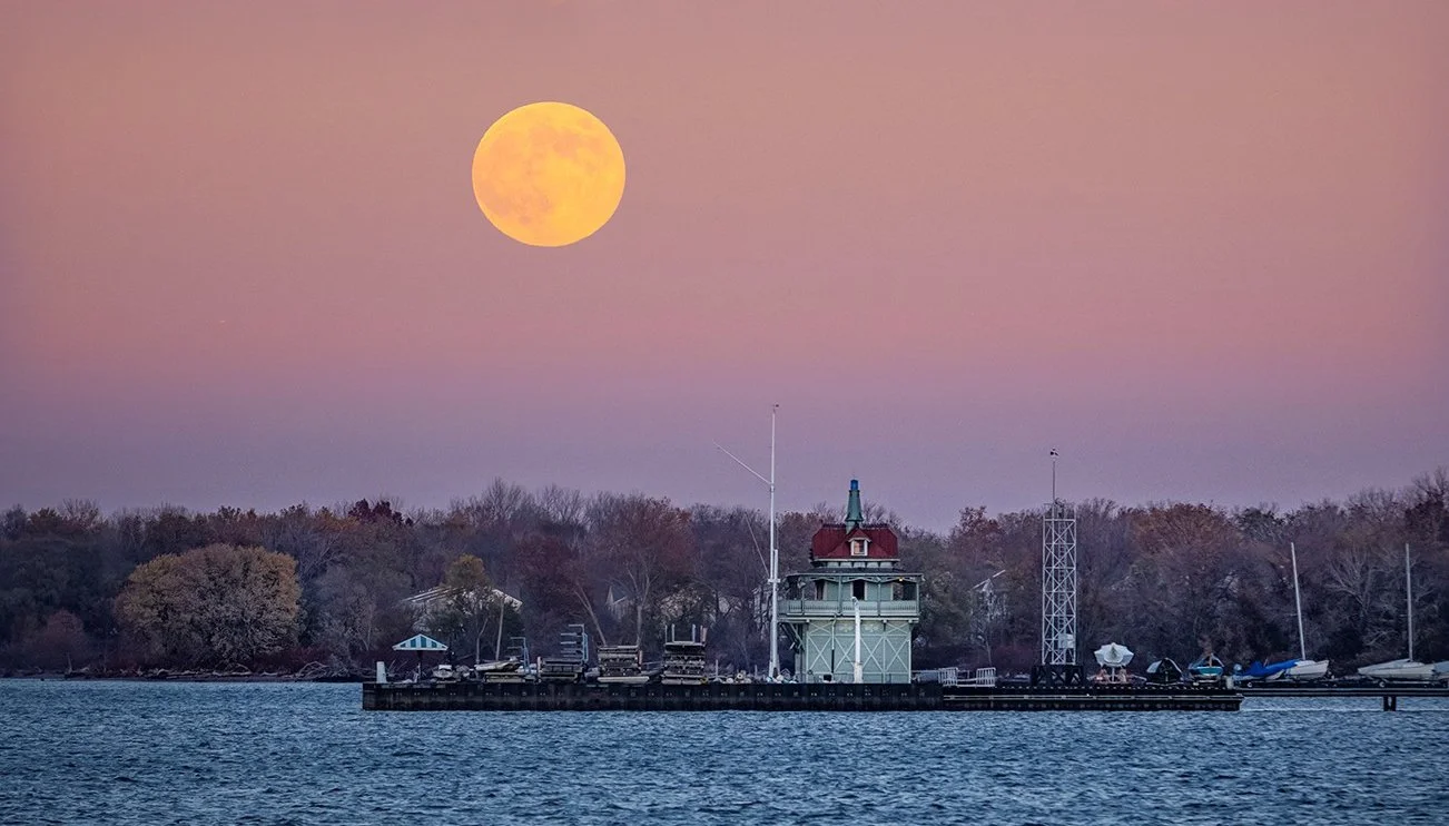 Full moon rising over the Delaware River in Palmyra with boats and Riverton Yacht Club on the shoreline, and trees in the background at dusk.