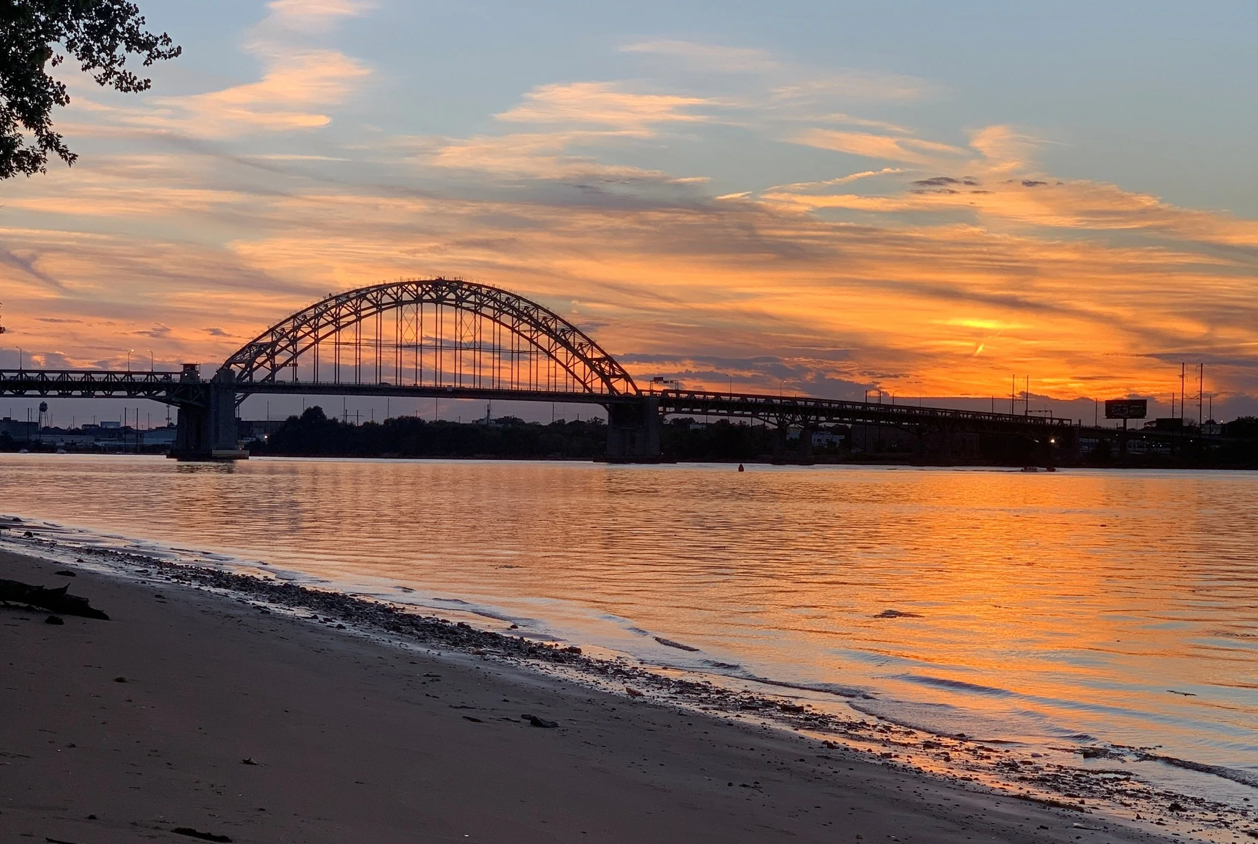 A sunset over a river with a bridge in the background. The sky is filled with orange and pink clouds, reflecting on the water. The scene includes a sandy shoreline with some debris.