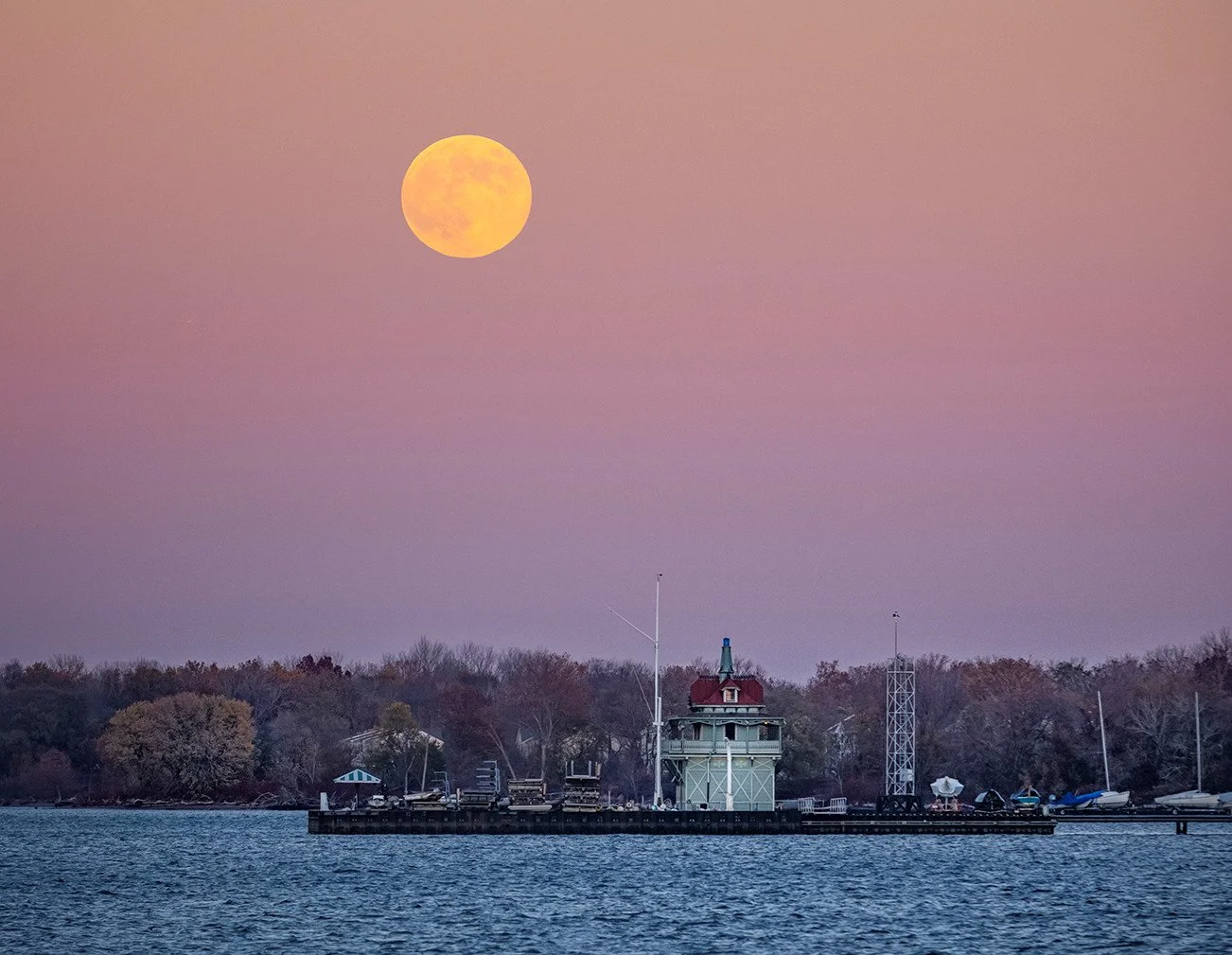 Full moon rising over a harbor with boats and a lighthouse against a pink and purple sky.