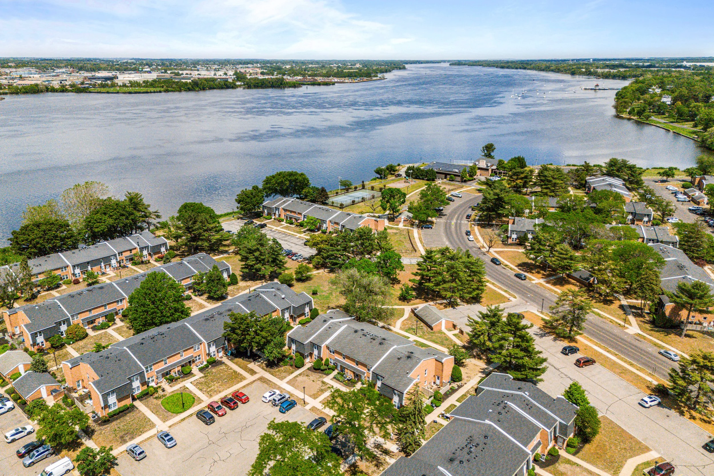 Aerial view of a residential area by a large river, with multiple apartment buildings, parking lots, and green trees under a partly cloudy sky.