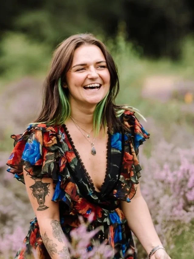 A woman with brown hair and green streaks, smiling and wearing a colorful floral dress with ruffles, standing outdoors in a field of purple flowers.  radio presenter, voice over Abi, female voice over artist, London voice over, United Kingdom