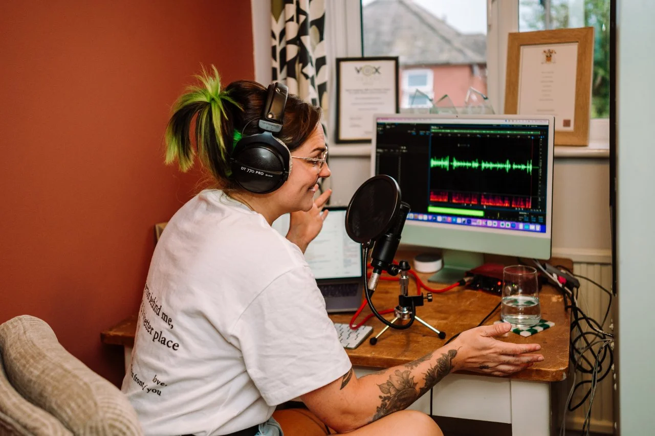 A woman with dyed green hair in a ponytail, wearing headphones, recording vocals at a microphone, with audio editing software on her desktop monitor. radio presenter, voice over Abi, female voice over artist, London voice over, United Kingdom