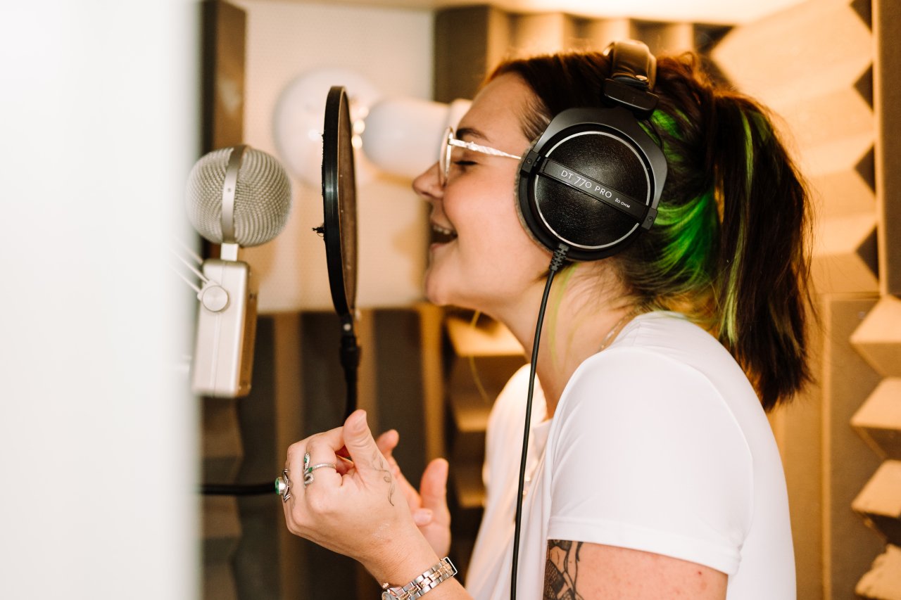 A woman in a recording studio wearing large headphones, singing into a microphone with a pop filter, wearing glasses, a white t-shirt, radio presenter, voice over Abi, female voice over artist, London voice over, United Kingdom