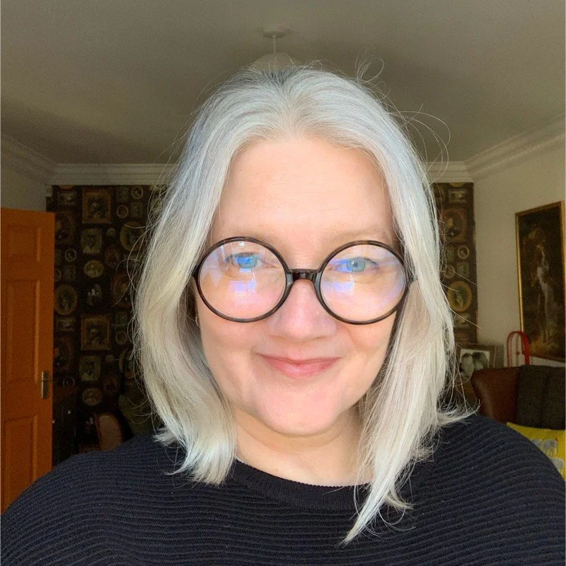 A woman with shoulder-length gray hair and round glasses taking a selfie indoors.