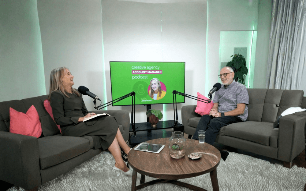 Two people sitting on sofas having a podcast interview, microphones in front of them, a woman on the left with a book, a man on the right with glasses, a large screen behind them displaying a podcast graphic with a woman, text reading "creative agency ACCOUNT MANAGER podcast hosted by JENNY PLANT."