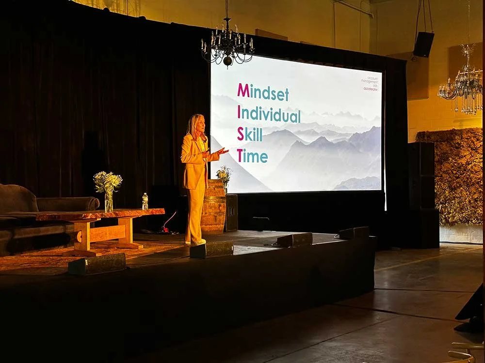 A woman is standing on a stage delivering a presentation. Beside her is a large screen displaying the words 'Mindset,' 'Individual,' 'Skill,' and 'Time' in pink and blue letters. The stage has a dark curtain backdrop, a small table with a vase of flowers, and some decorative items. The setting appears to be an indoor conference or event space with chandeliers hanging from the ceiling.