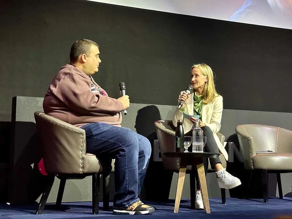 Two women sitting on stage in armchairs, engaged in a conversation, each holding a microphone; one woman is wearing a brown jacket and jeans, and the other is wearing a white blazer and black pants, with a table in between holding water bottles, glasses, and a notebook.