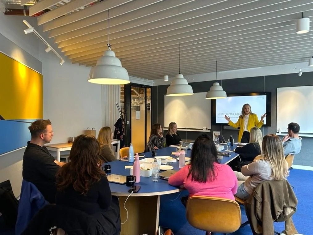 A woman in a yellow blazer giving a presentation to a group of people seated around a blue conference table in a modern office meeting room. There is a large screen behind her displaying a presentation slide.