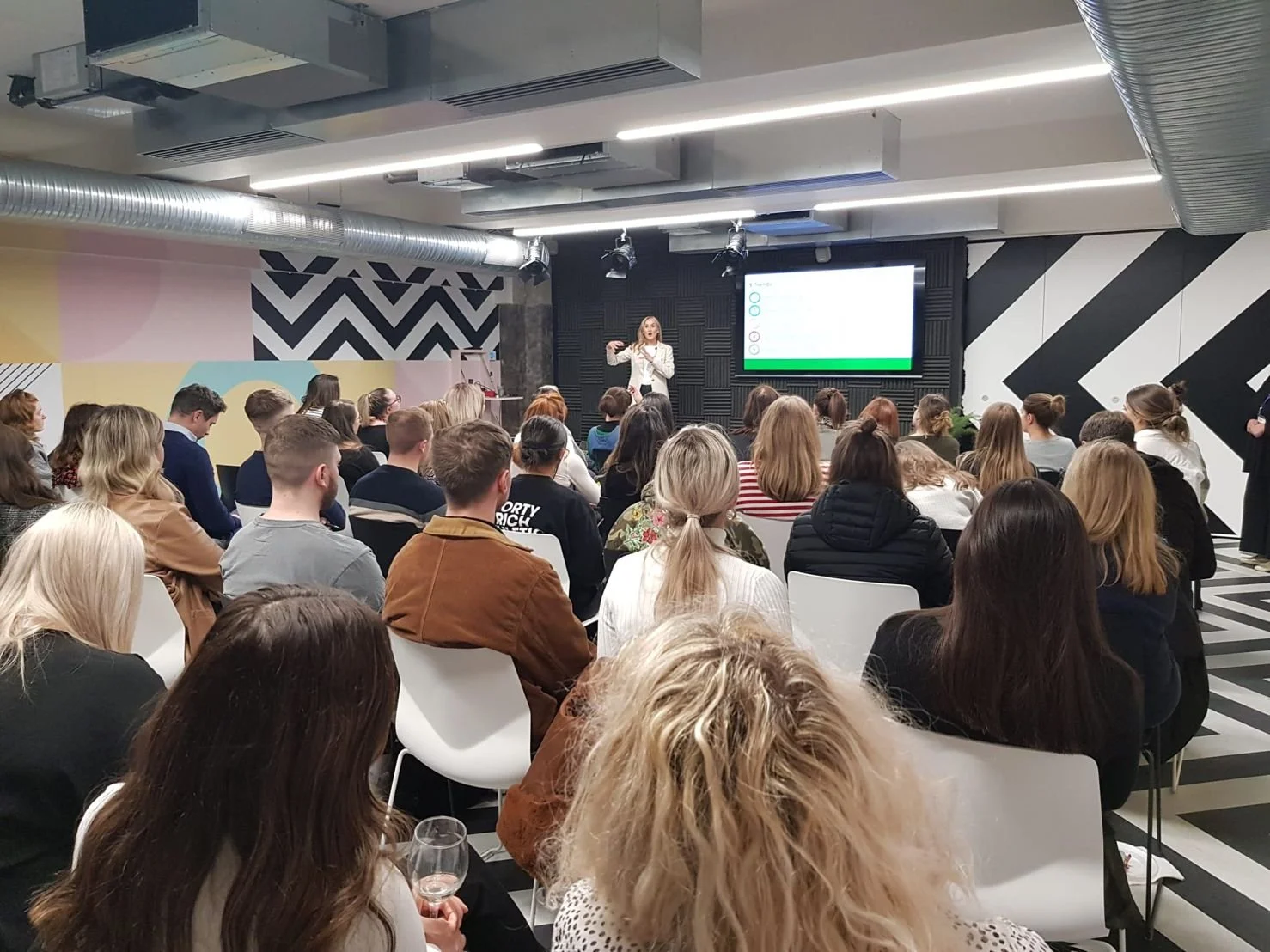 A woman standing on a stage giving a presentation to an audience in a modern conference room with black and white geometrical wall patterns.