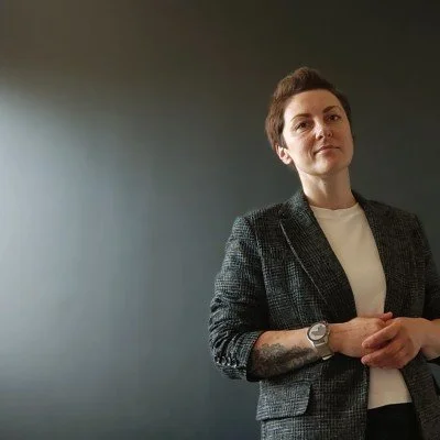 A lady with short hair, wearing a grey suit with a white top.