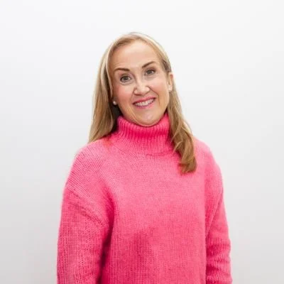 A woman with blonde hair wearing a pink turtleneck sweater smiling against a plain white background.