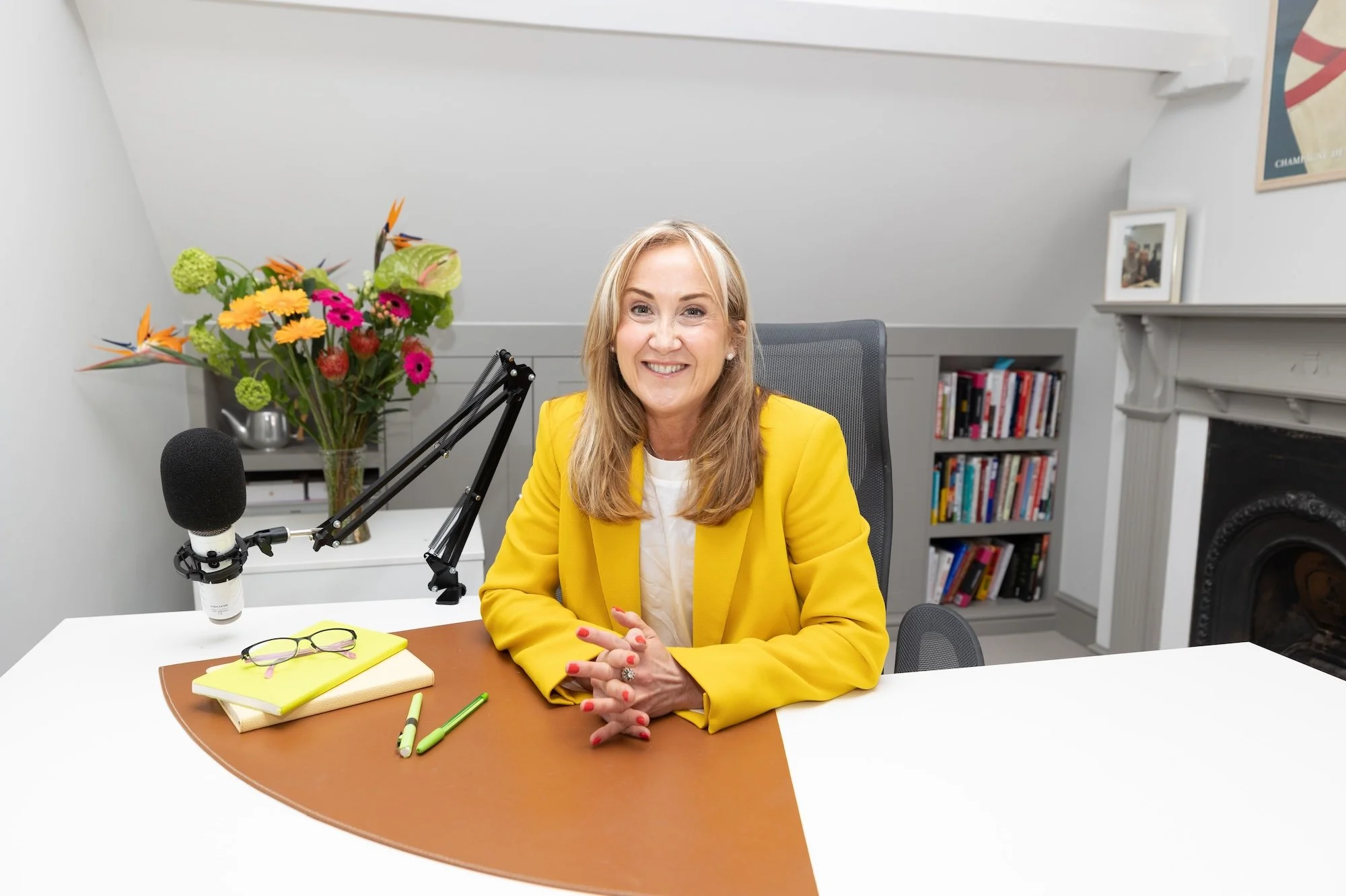 A woman sitting at a desk in a bright room, smiling, with a bouquet of colorful flowers, a microphone, notebooks, and pens on the desk, and a bookcase in the background.
